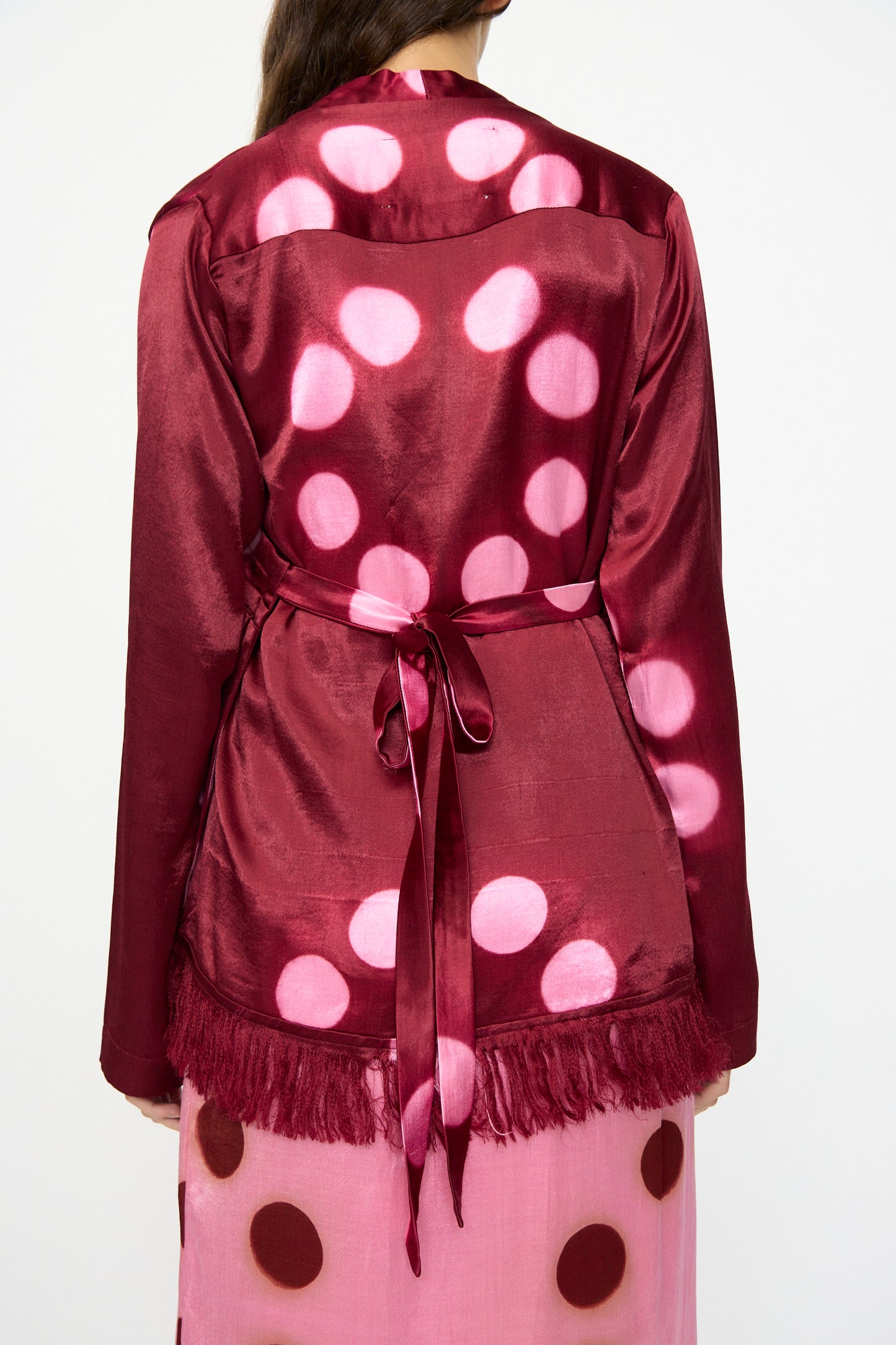 A person is shown from the back wearing the TIGRA TIGRA Silk Mashroo Moon Dye Wrap Top in Maroon, featuring a shiny finish, large pink polka dots, a matching belt, and fringe hem, styled over a pink skirt with brown spots.
