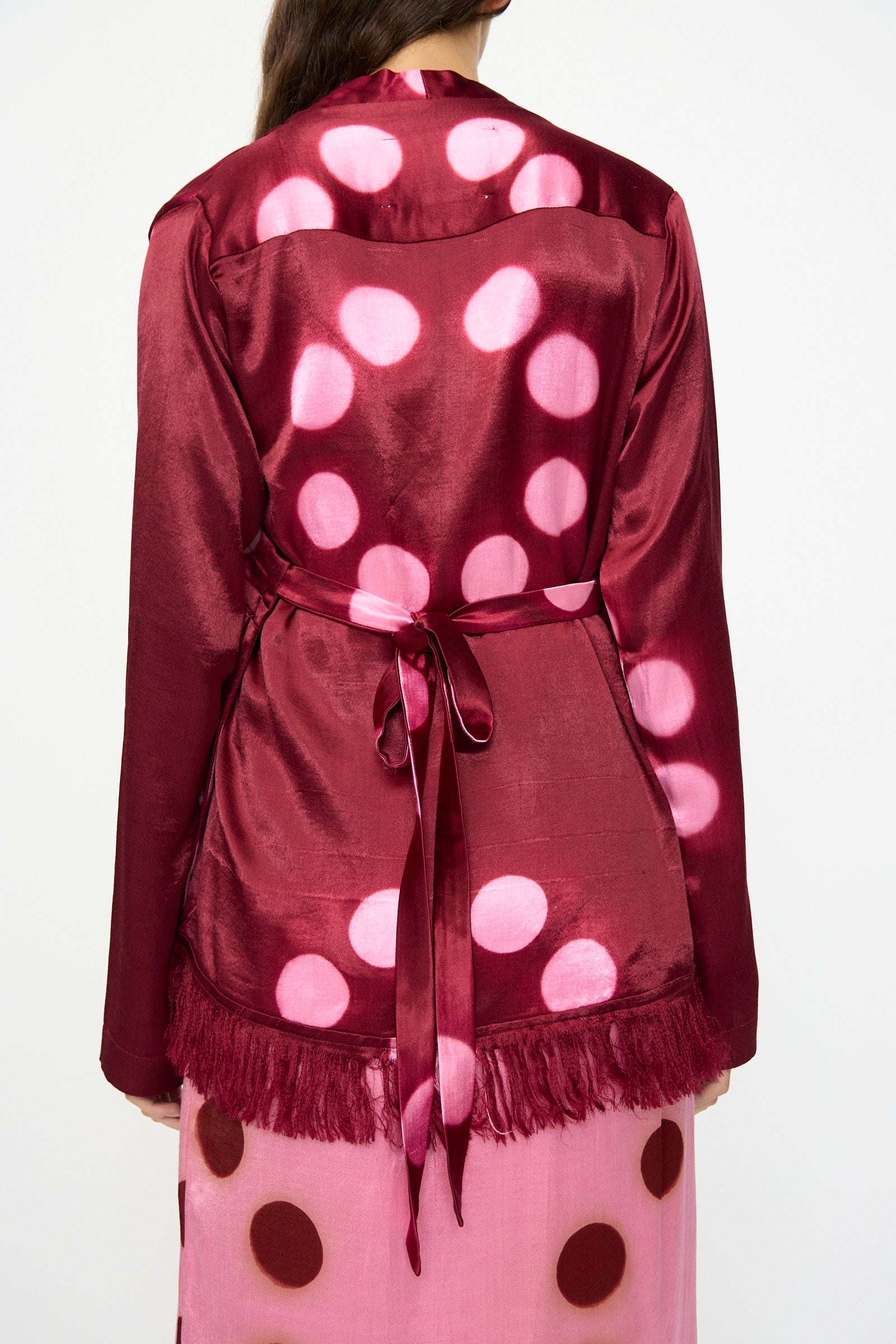 A person is shown from the back wearing the TIGRA TIGRA Silk Mashroo Moon Dye Wrap Top in Maroon, featuring a shiny finish, large pink polka dots, a matching belt, and fringe hem, styled over a pink skirt with brown spots.