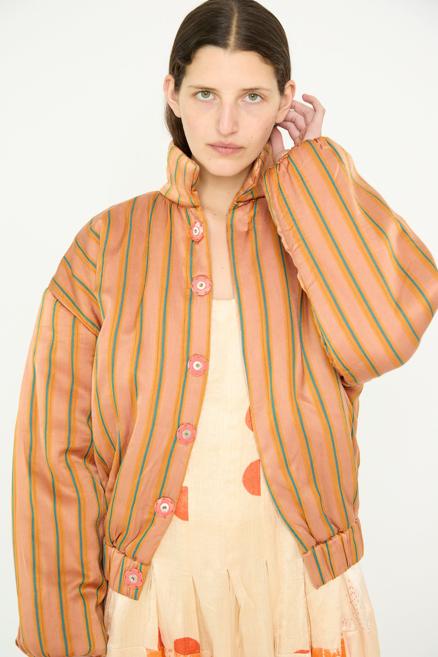 A model with long hair is wearing a TIGRA TIGRA Silk Mashroo Striped Aviator Puff Jacket in Peach and a dress with orange dots, standing against a plain background with one arm raised. 