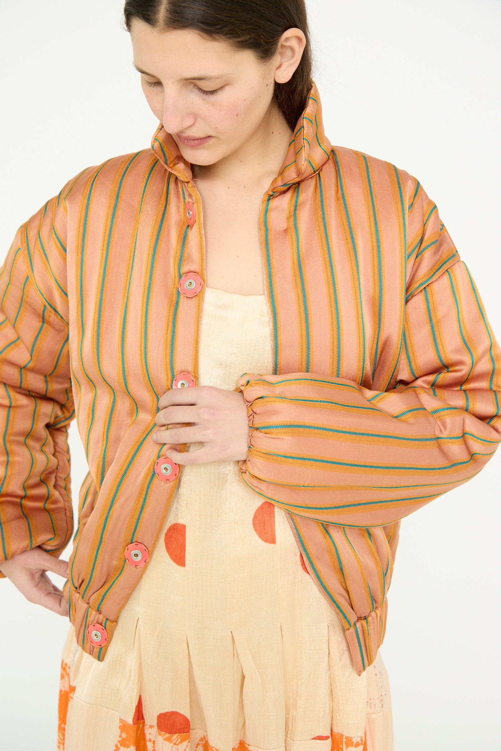 A model wearing the TIGRA TIGRA Silk Mashroo Striped Aviator Puff Jacket in Peach over a beige dress with orange spots, looking down against a white backdrop. 