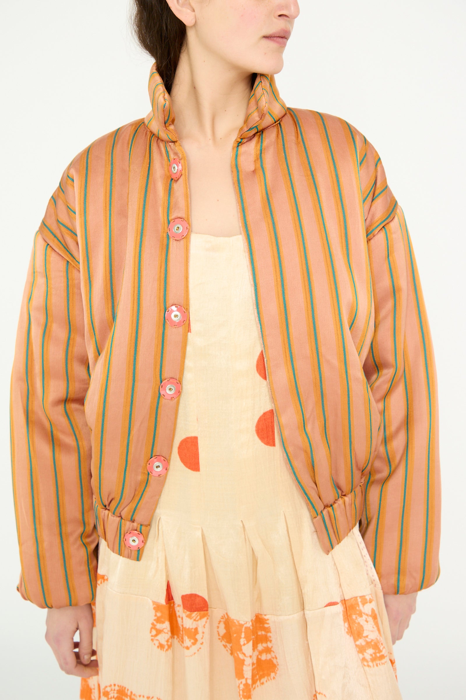 A model is in the TIGRA TIGRA Silk Mashroo Striped Aviator Puff Jacket in Peach over a beige dress with orange patterns, against a white backdrop. 