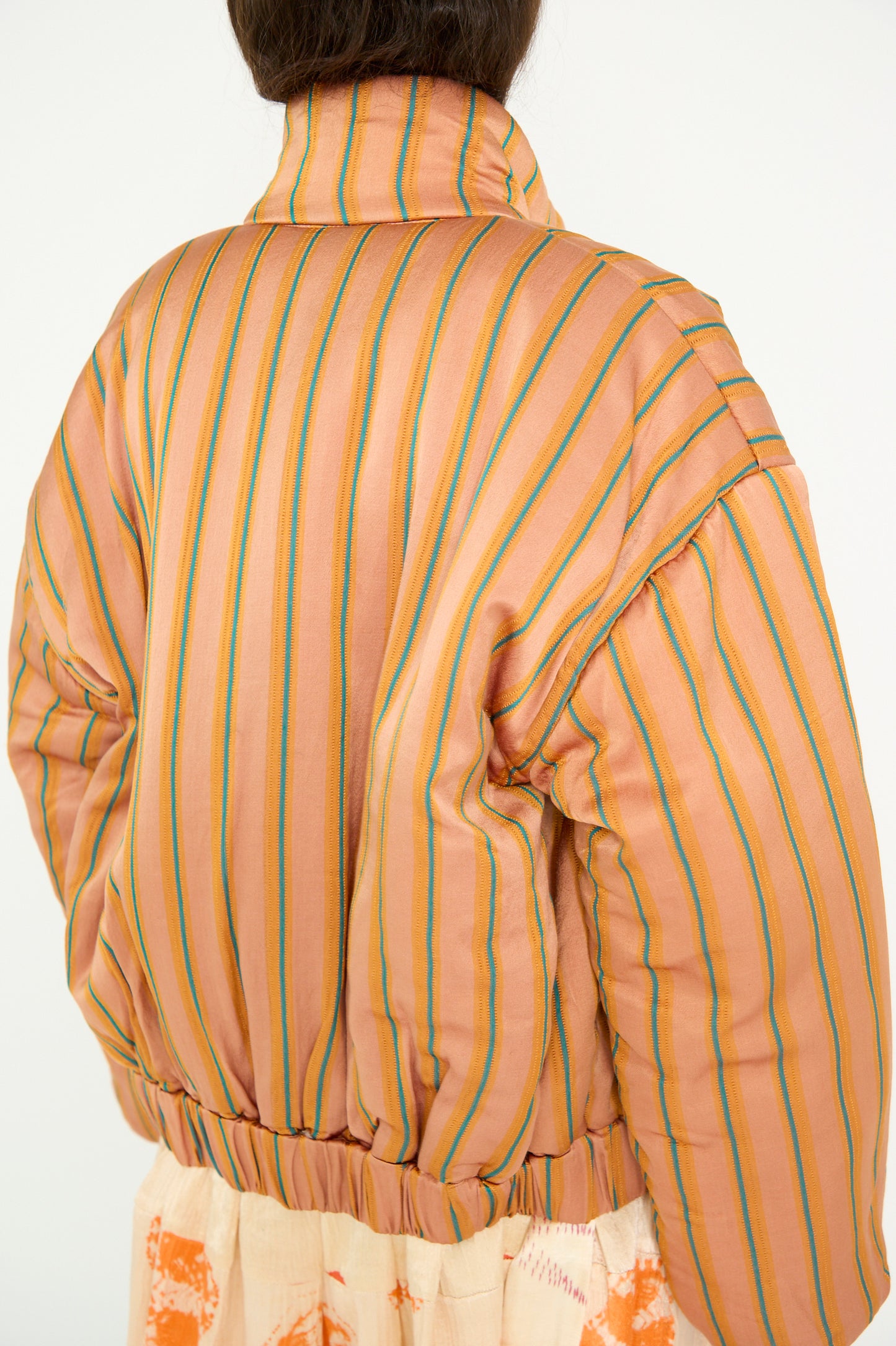 A person is shown from the back wearing a TIGRA TIGRA Silk Mashroo Striped Aviator Puff Jacket in Peach with orange, yellow, blue, and peach stripes, against a white backdrop. 