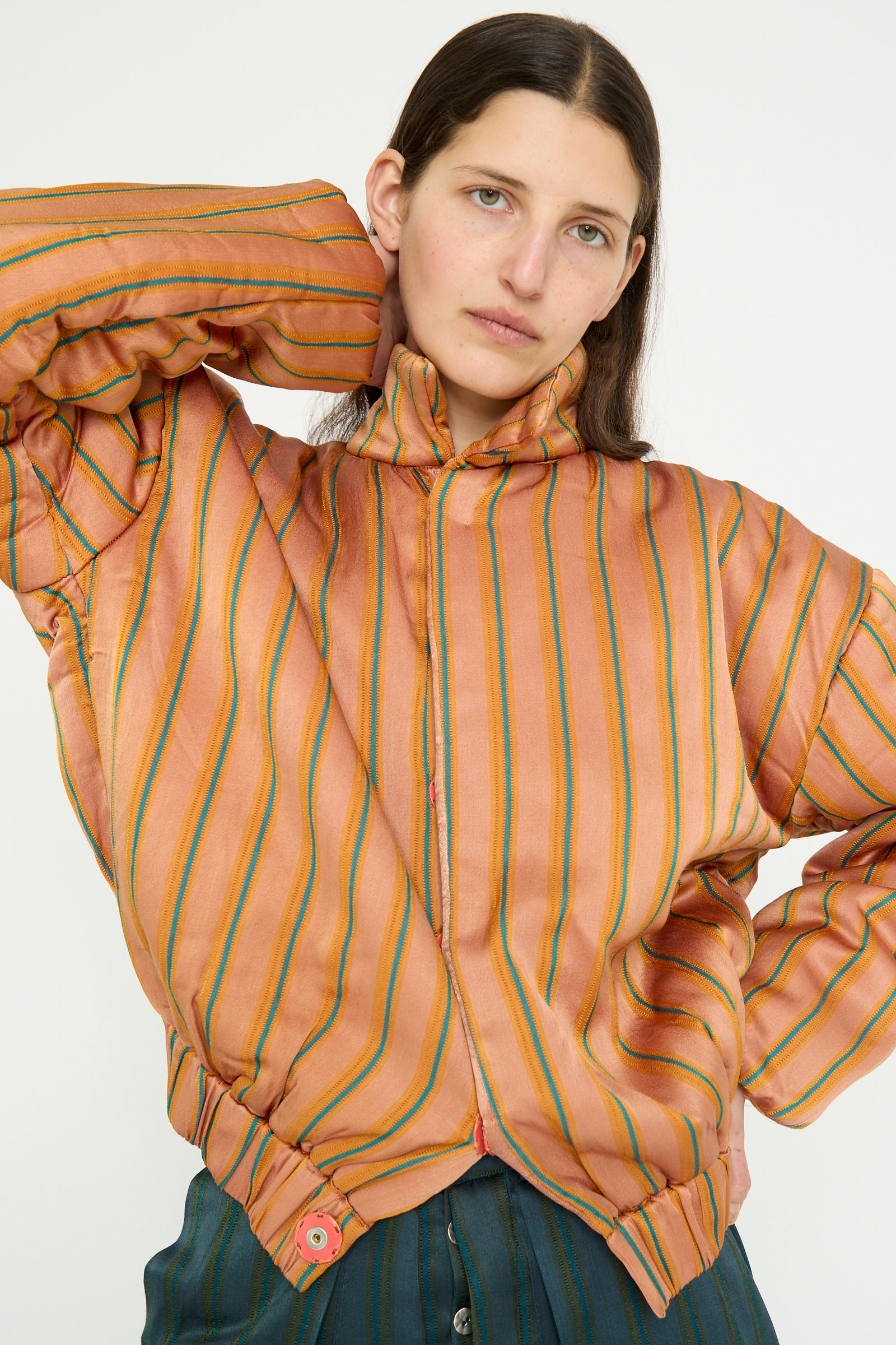 A person rests one hand on their head while wearing the oversized Silk Mashroo Striped Aviator Puff Jacket in Peach by TIGRA TIGRA, looking forward against a white backdrop. 