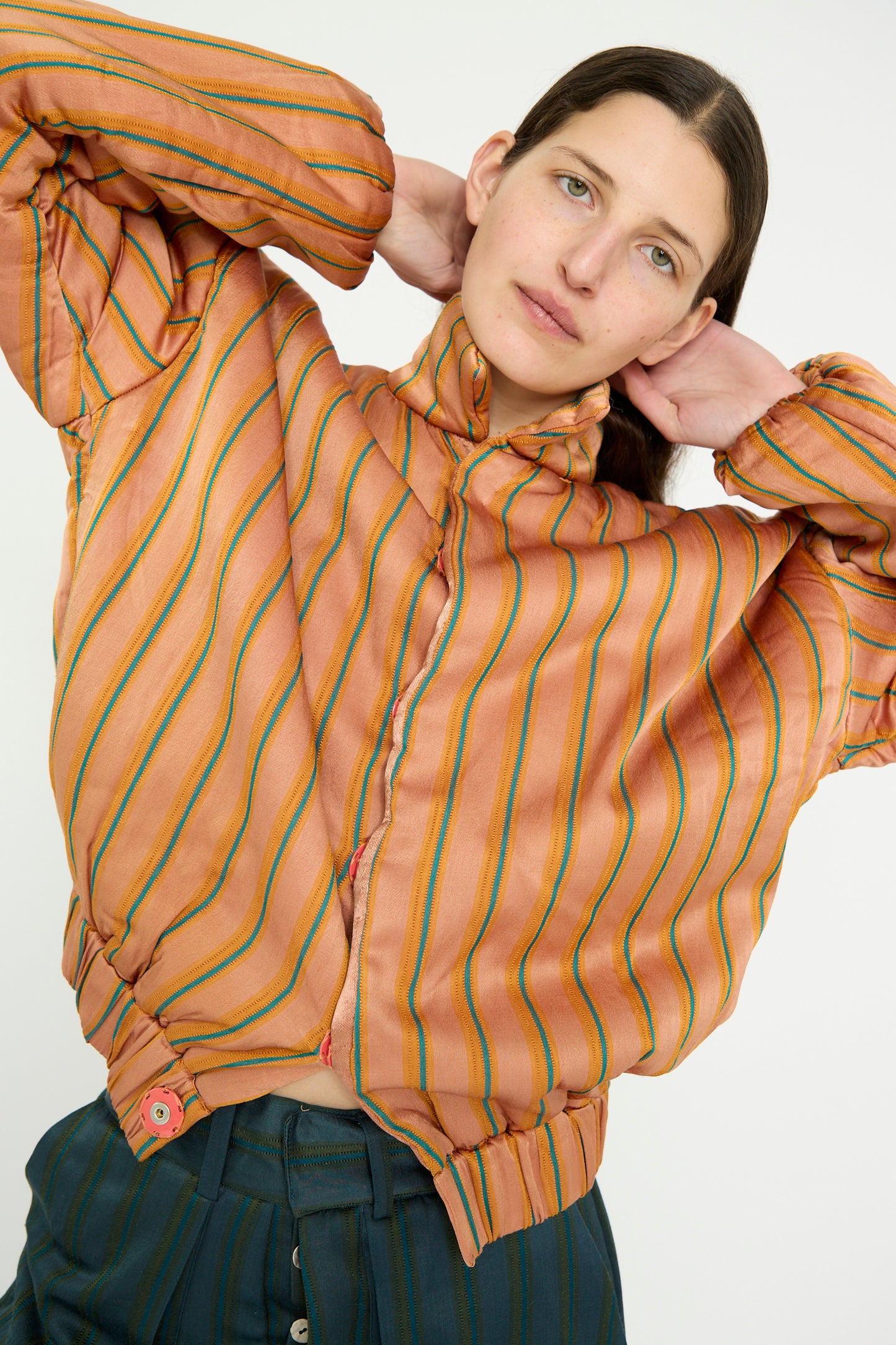 A model in the TIGRA TIGRA Silk Mashroo Striped Aviator Puff Jacket in Peach poses with their hands behind their head against a white backdrop. 