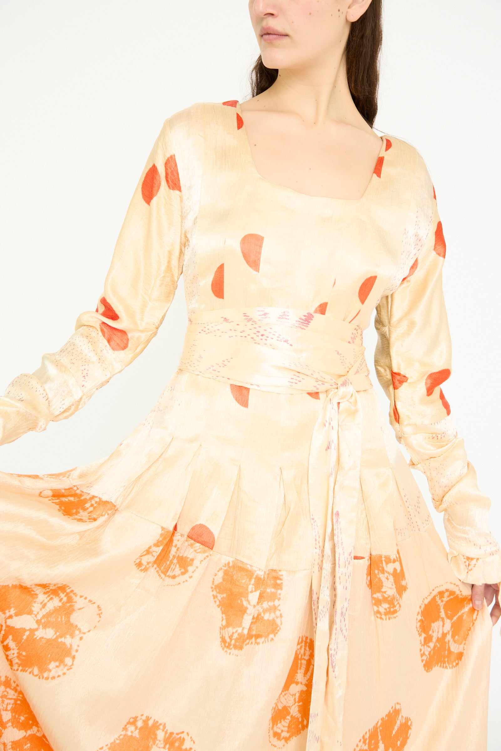 A person models the TIGRA TIGRA Tropical Dress in pale grey with orange Bandhej and long sleeves, accentuated by a waist tie, set against a plain background.