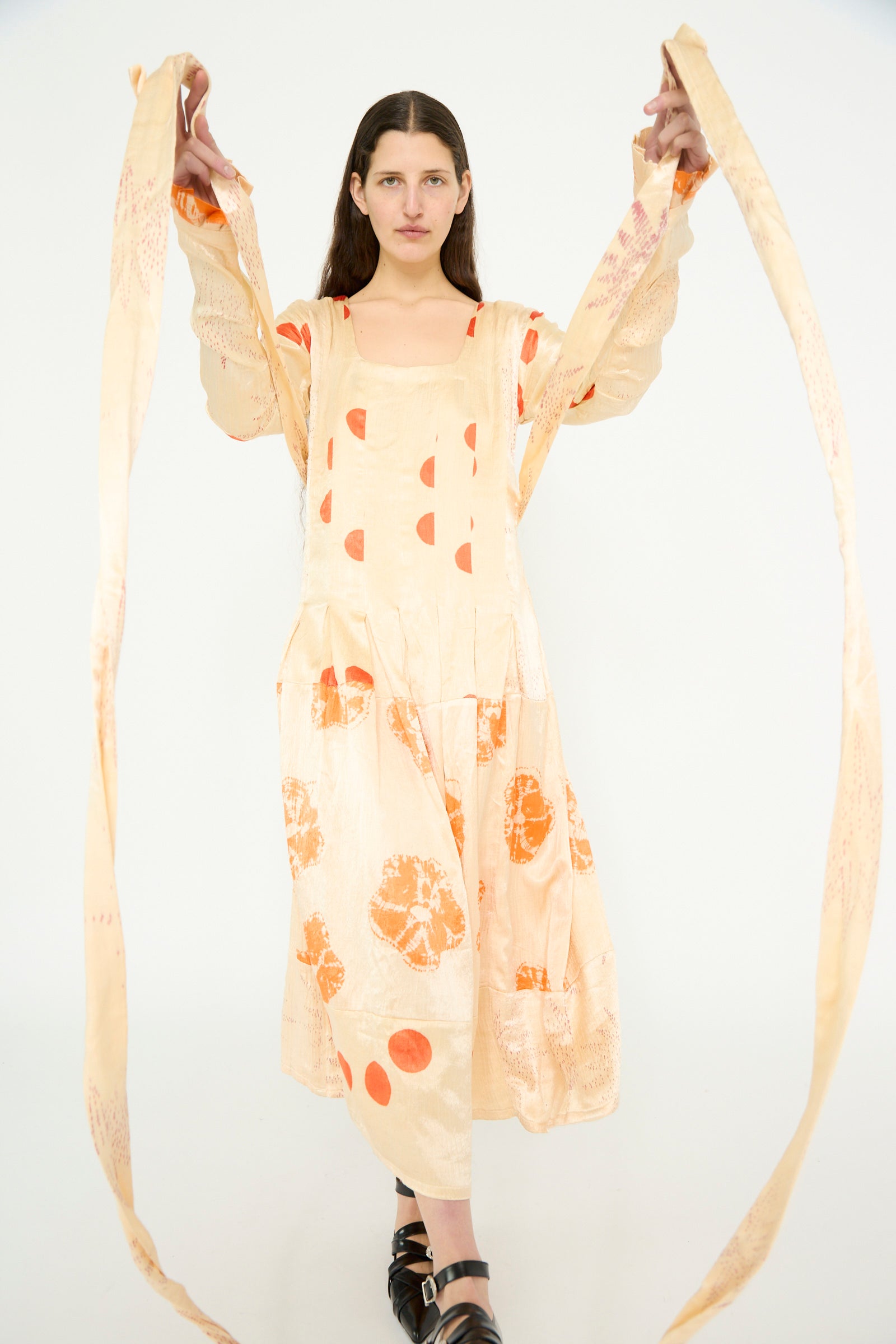 A model poses in a TIGRA TIGRA Tropical Dress featuring patchwork tie dye in cream with orange bandhej patterns, showcasing its long self-tie belt, against a white backdrop. 