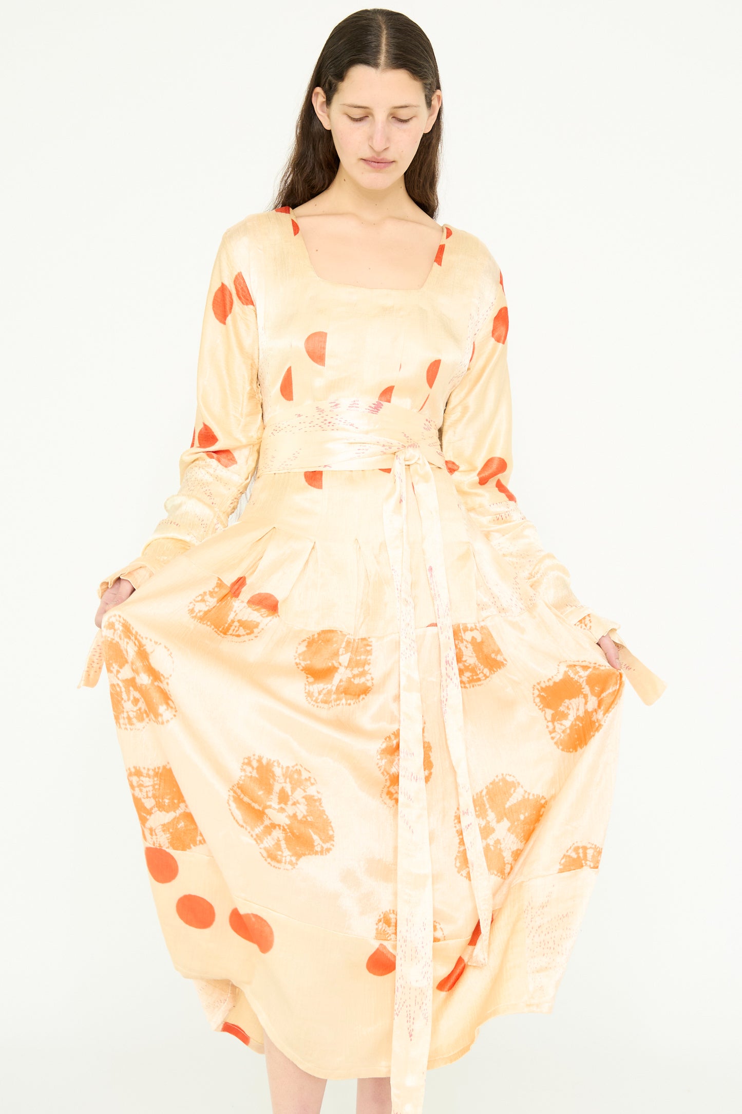 A model looks down while holding the sides of their cream maxi dress, adorned with a patchwork tie-dye pattern in pale grey and orange bandhej by TIGRA TIGRA, against a white backdrop.