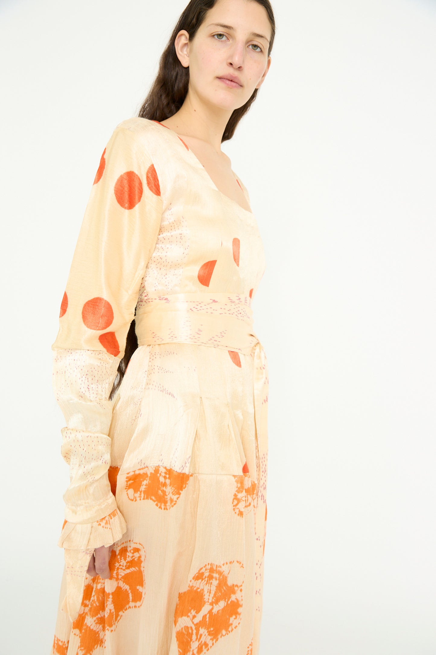 A person stands against a plain background wearing the Tropical Dress by TIGRA TIGRA, featuring long sleeves and patchwork tie-dye in pale grey with orange bandhej patterns.