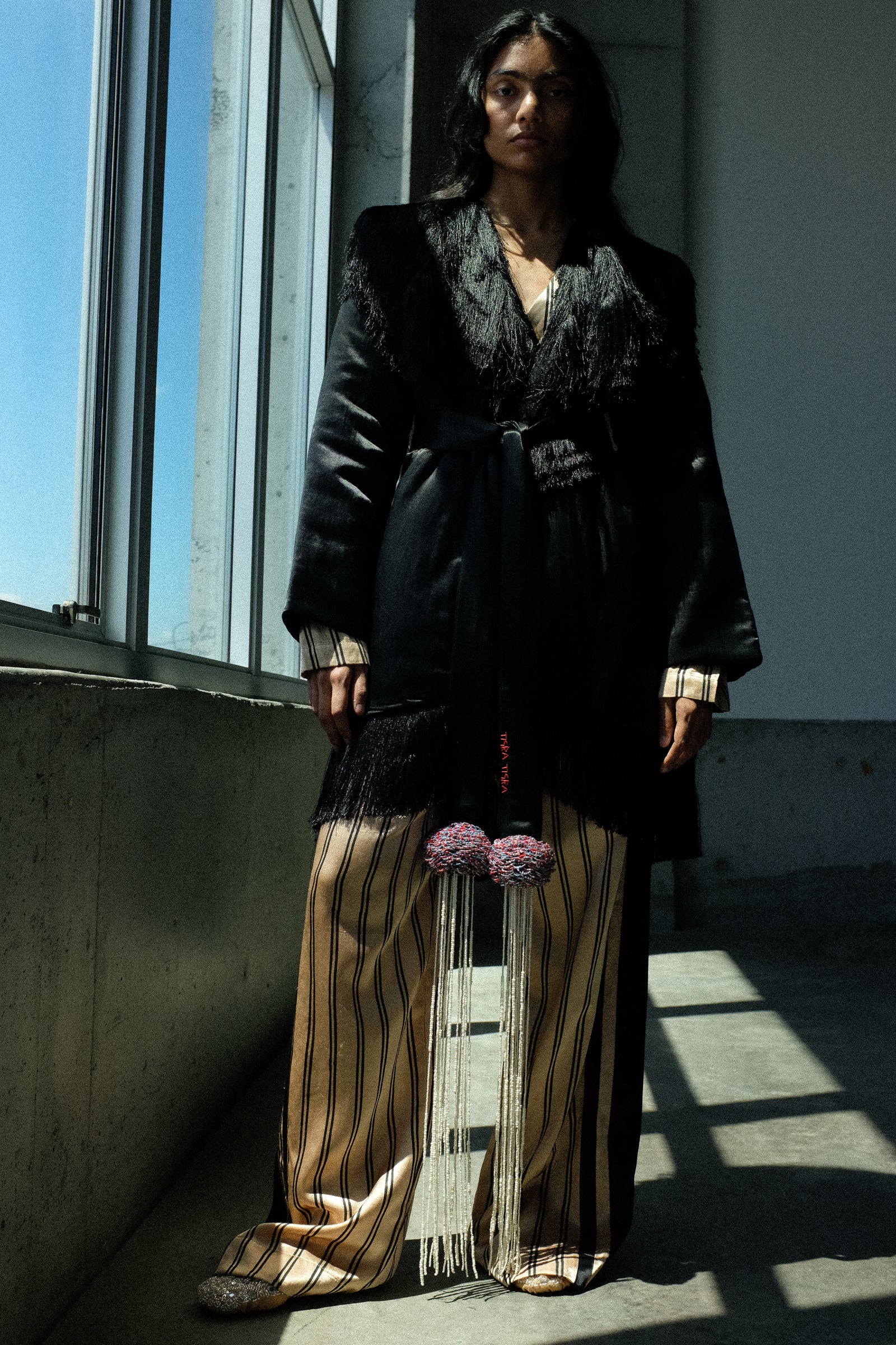 A model stands in a sunlit industrial room wearing a black wrap-style coat with dark textured trim over a striped beige and brown tunic, paired with wide-leg striped trousers and a long fringe tassel accessory.