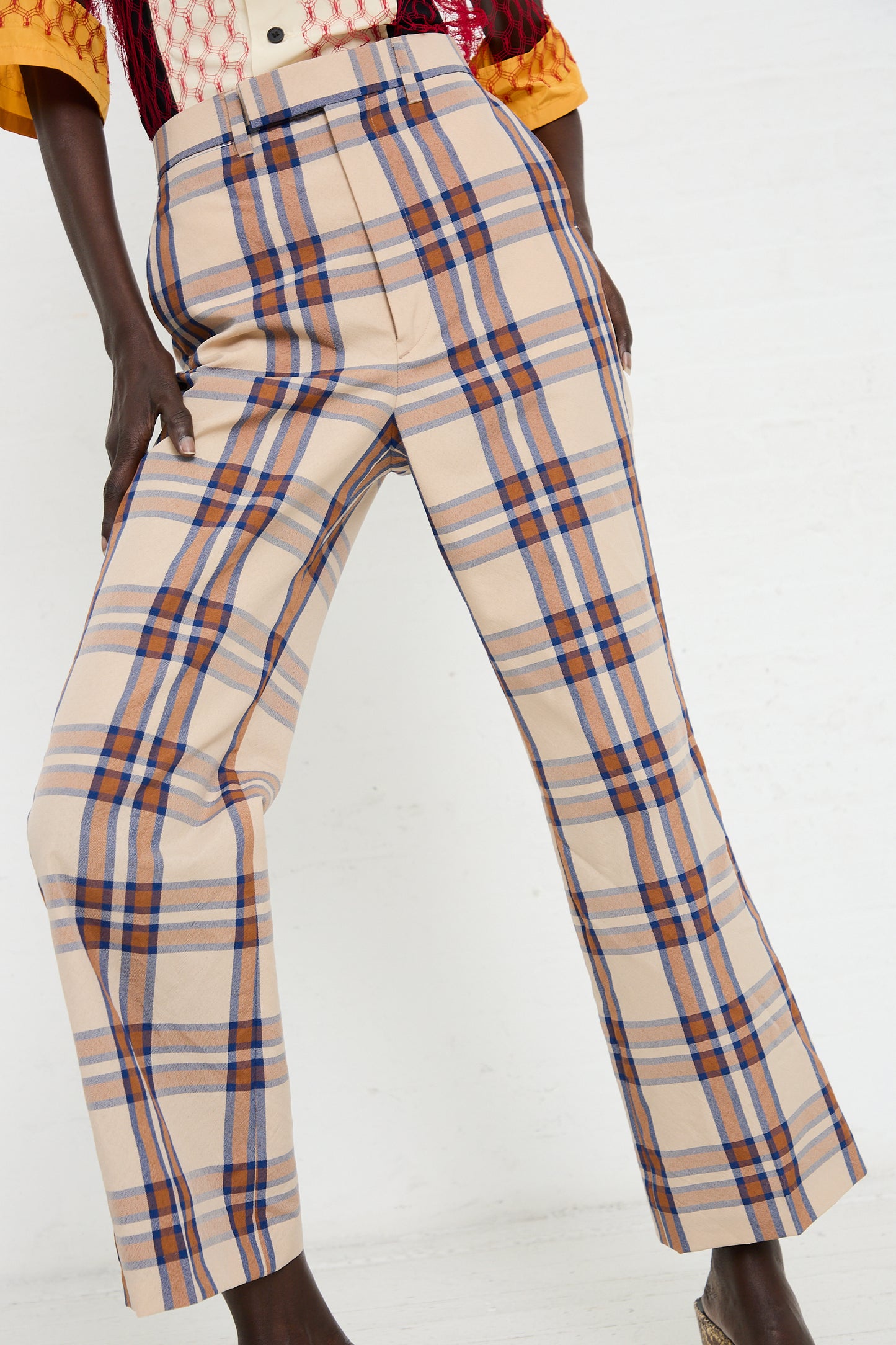 A person stands against a plain background, showcasing the TOGA ARCHIVES Wool Check Pant in Beige with a blue and red wool check plaid pattern. The lower body is visible in high-waisted trousers with slightly flared legs.