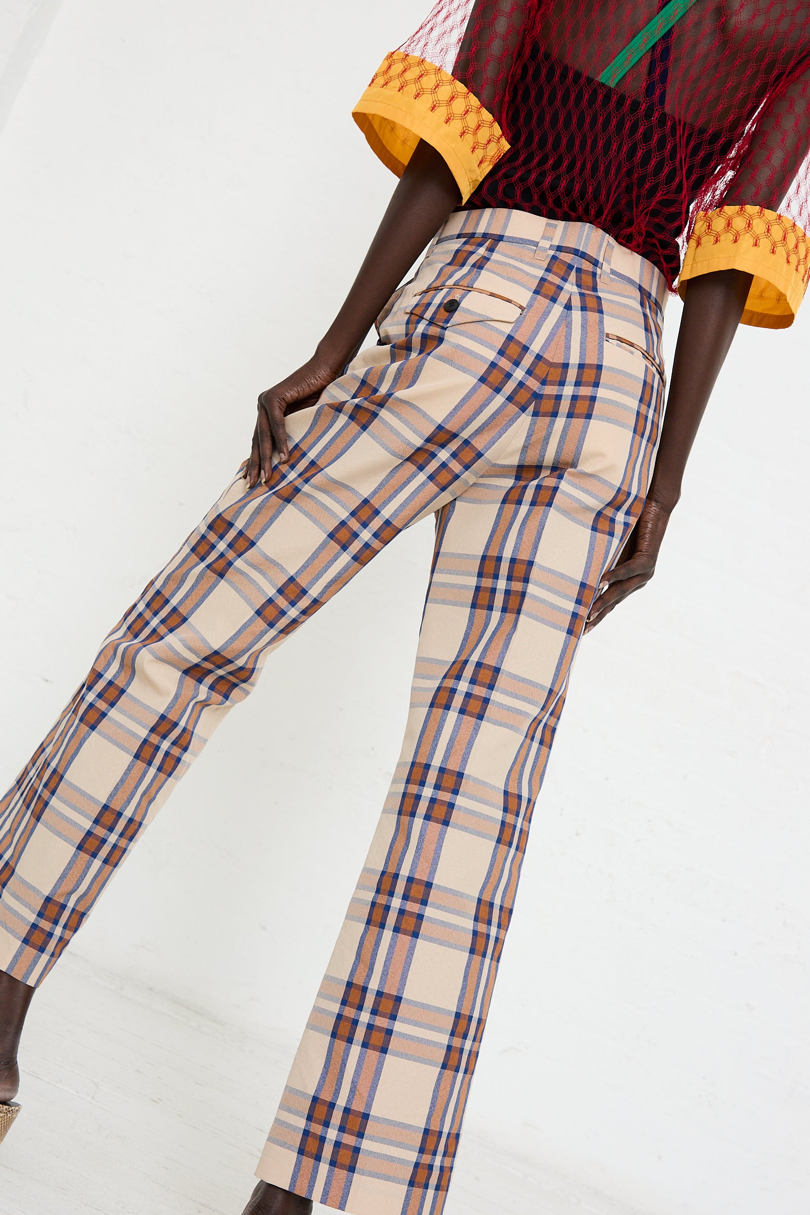 A person stands sideways wearing TOGA ARCHIVES Wool Check Pant in Beige with slightly flared legs and hands on hips, showcasing the rear and side view of the pants. The person also wears a colorful, textured top.