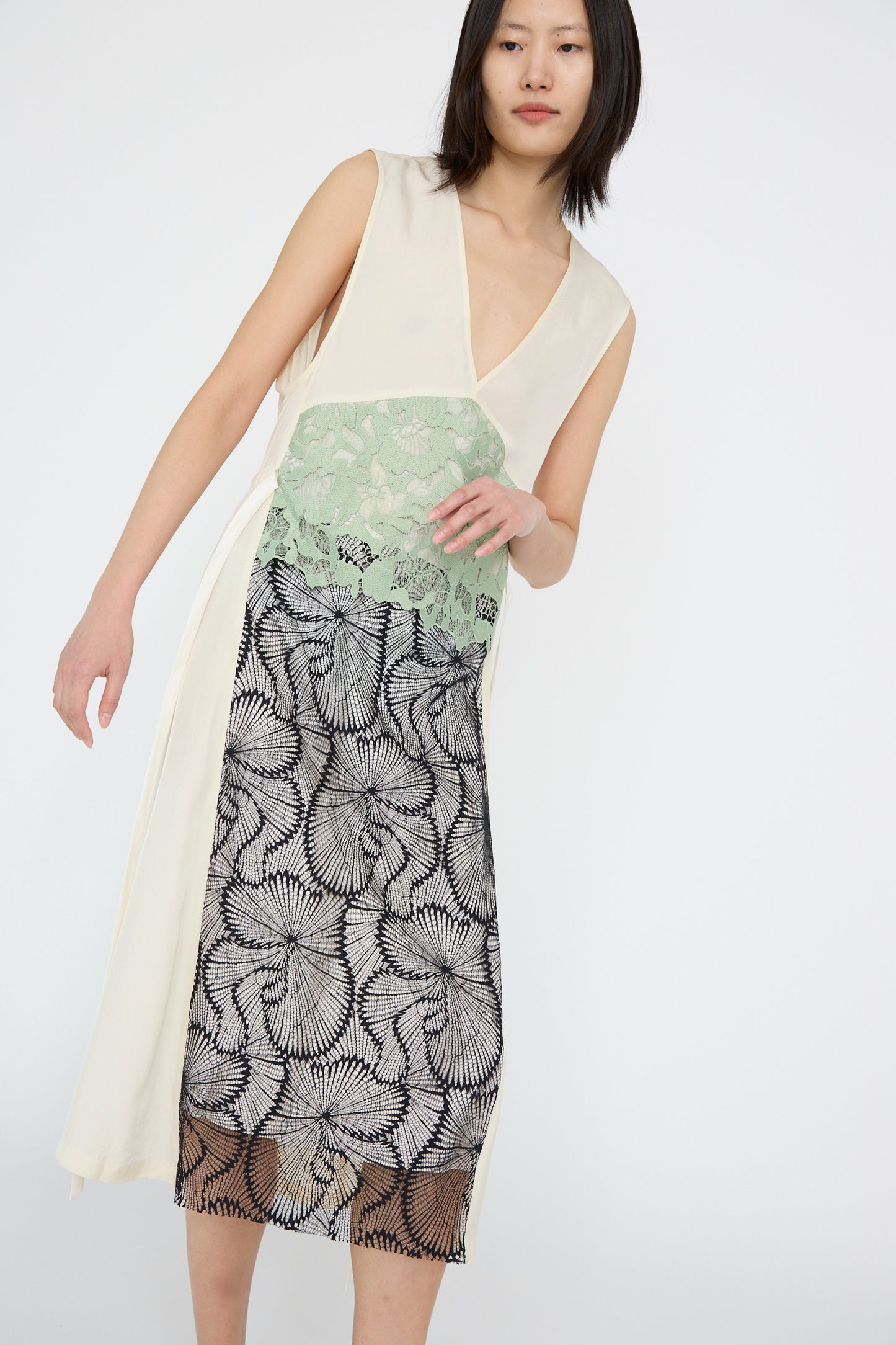 A person wearing the TOGA ARCHIVES Acetate Twill Lace Dress in Off White, featuring a white V-neck top, green lace middle, and black patterned skirt, stands against a plain background.