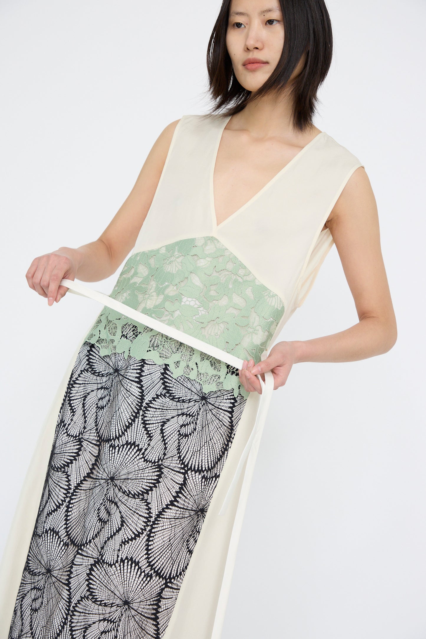 Wearing the TOGA ARCHIVES Acetate Twill Lace Dress in Off White, the model adjusts their white belt, wearing a sleeveless maxi dress with a deep V-neckline, green floral pattern, and black/white abstract design