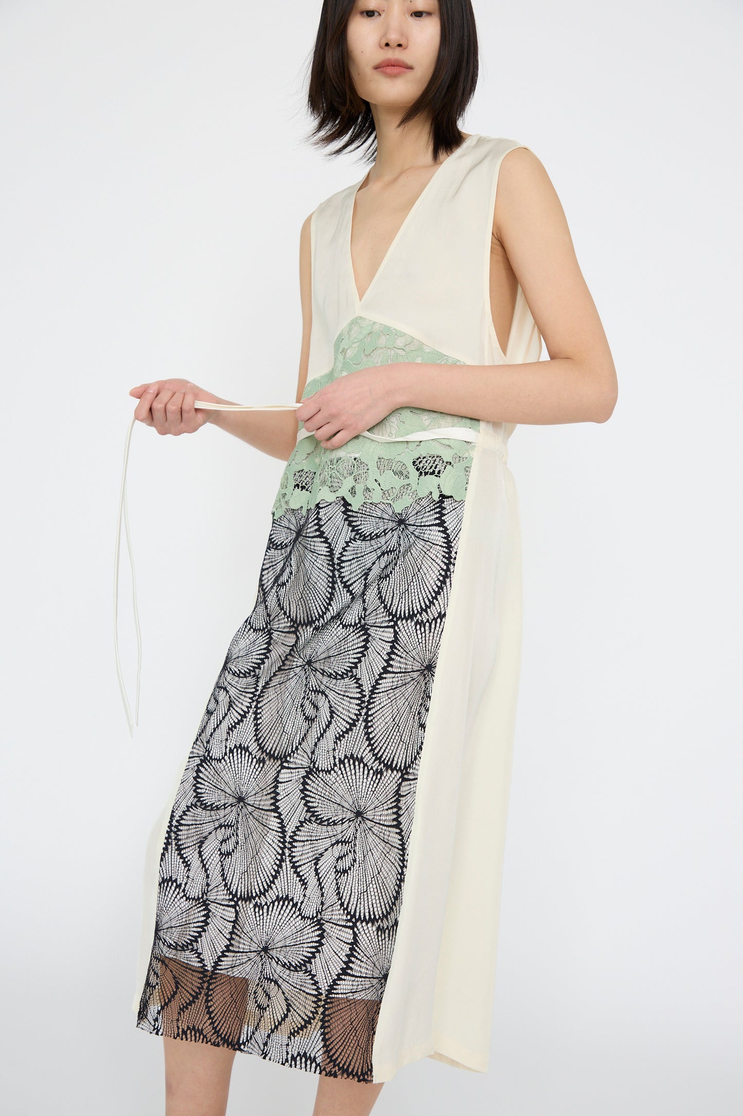 A model wears a sleeveless Acetate Twill Lace Dress by TOGA ARCHIVES, featuring a deep V-neck and showcasing green lace over black floral patterns. She holds a string in her hands against a white background.