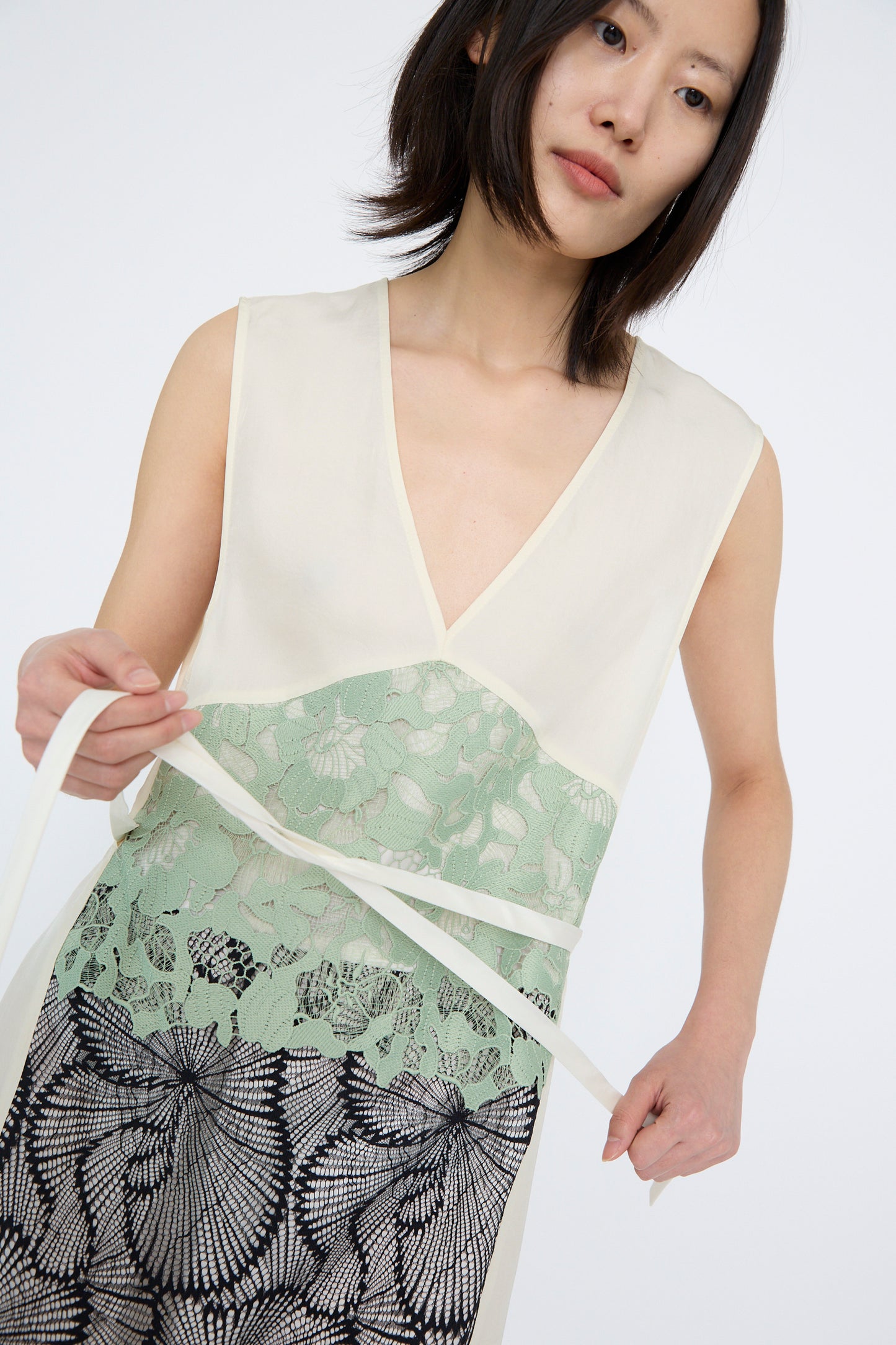 A woman wears the TOGA ARCHIVES Acetate Twill Lace Dress in Off White, featuring a V-neck design and green and black lace patterns. She ties a white belt around her waist, against a white background. 