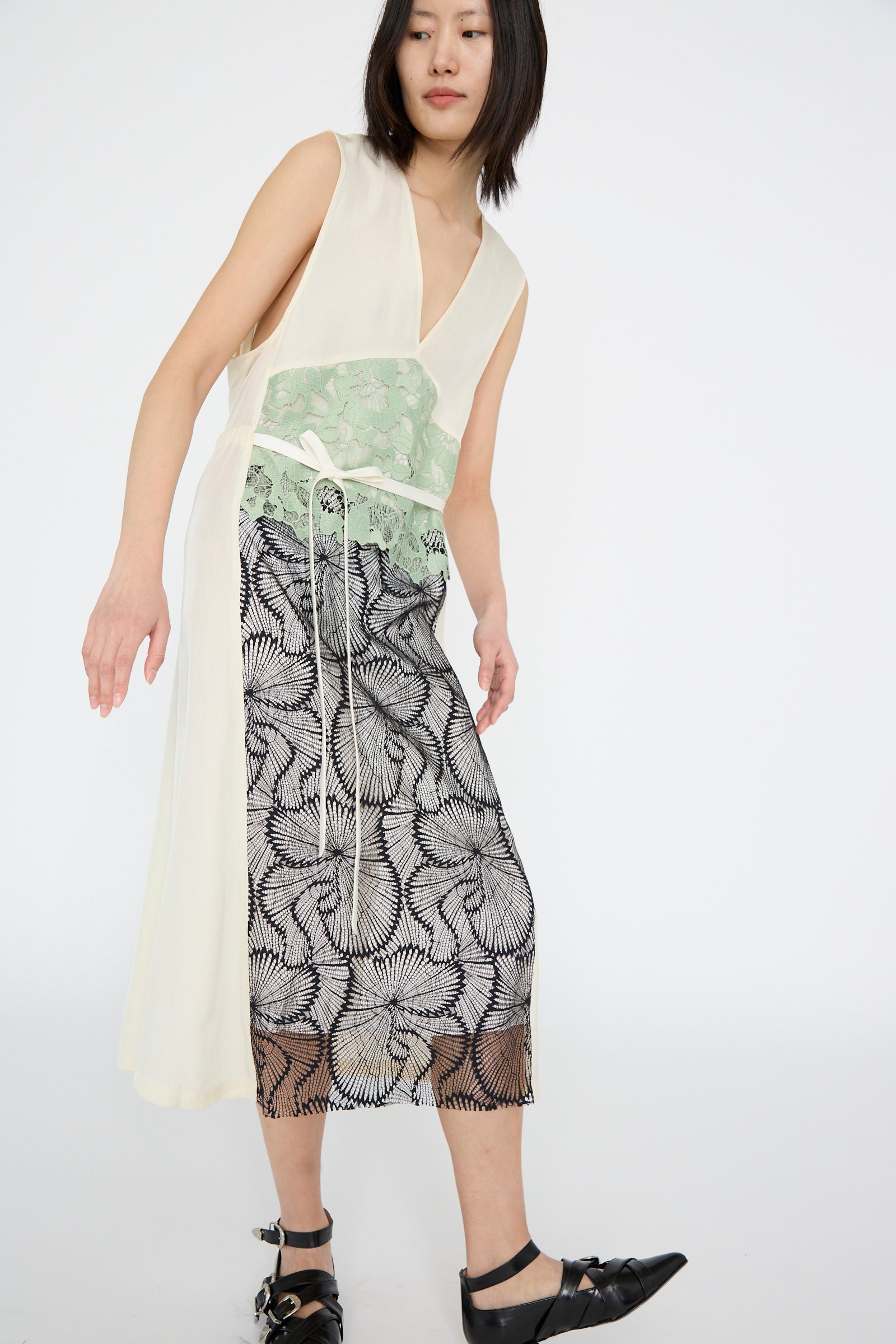 A woman stands against a plain background in an Acetate Twill Lace Dress by TOGA ARCHIVES, featuring a sleeveless green lace bodice and black patterned skirt. 