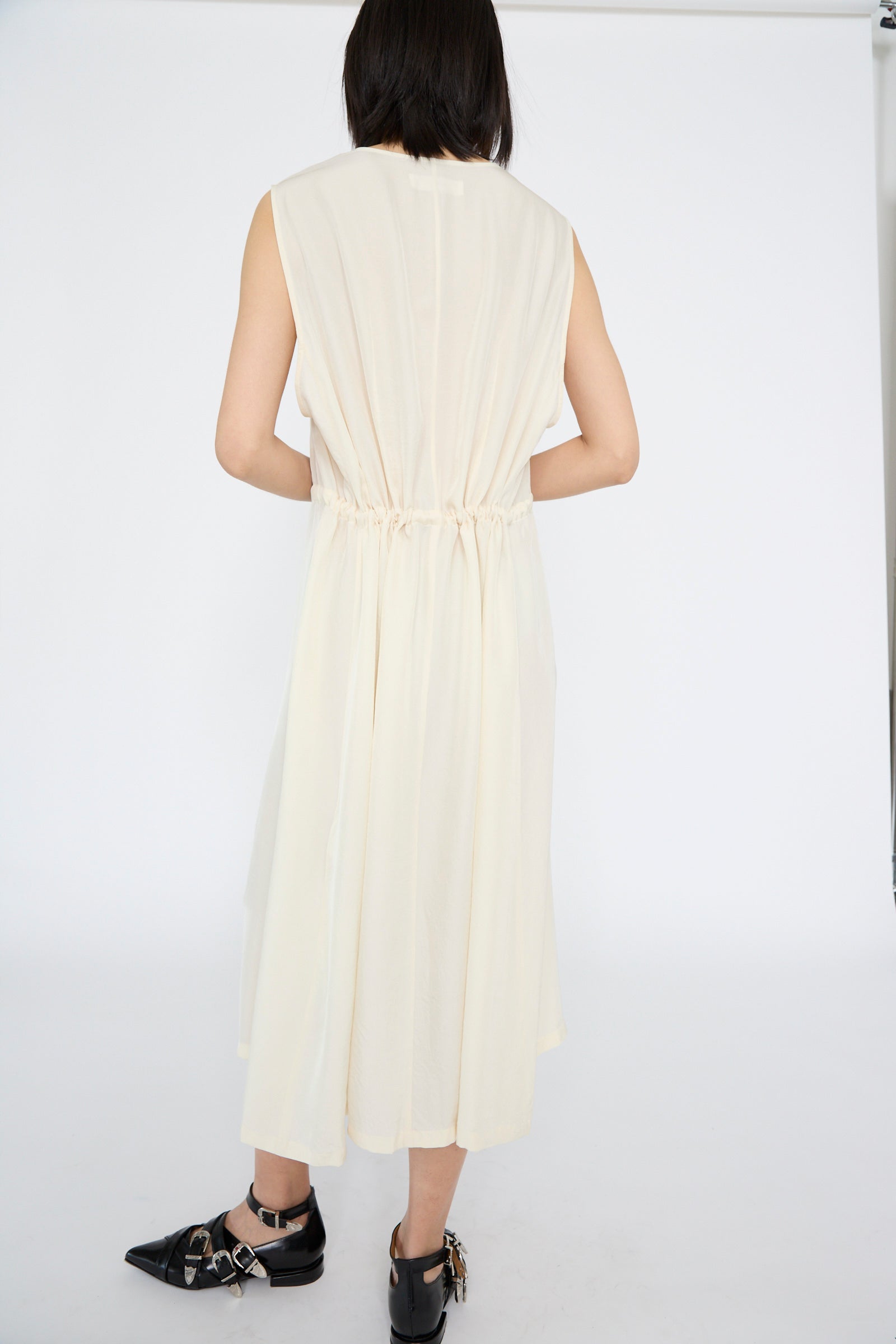 Wearing the TOGA ARCHIVES Acetate Twill Lace Dress in Off White, a person stands with their back to the camera. The dress features a sleeveless design with a cinched waist and V-neck. They pair it with black pointed shoes adorned with silver embellishments.