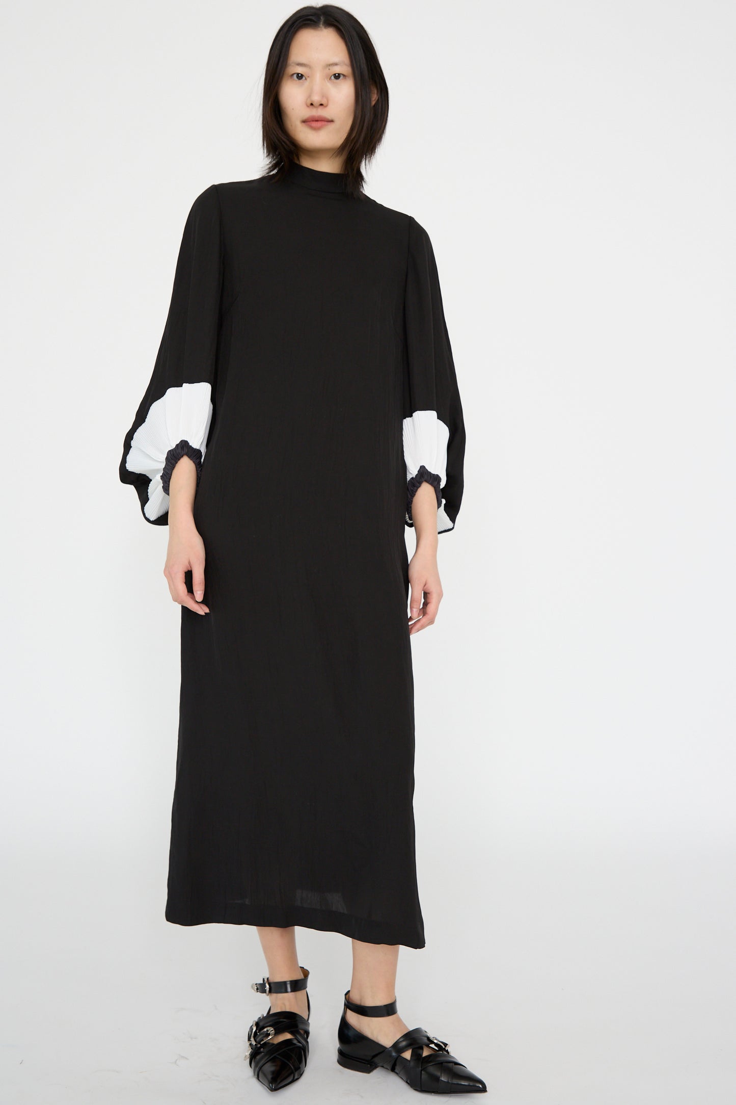 A person wears the Acetate Twill Pleats Dress in Black by TOGA ARCHIVES, showcasing bishop sleeves with white accents, paired with black shoes against a plain white background.