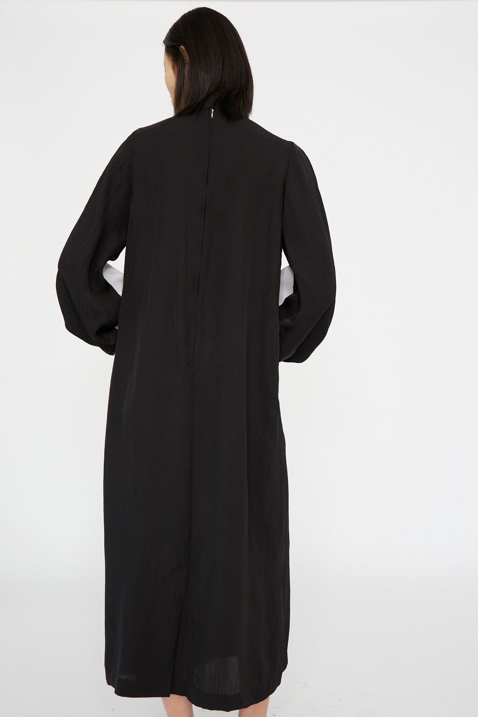 A person with dark hair is shown from the back, wearing a TOGA ARCHIVES Acetate Twill Pleats Dress in Black, set against a plain background.