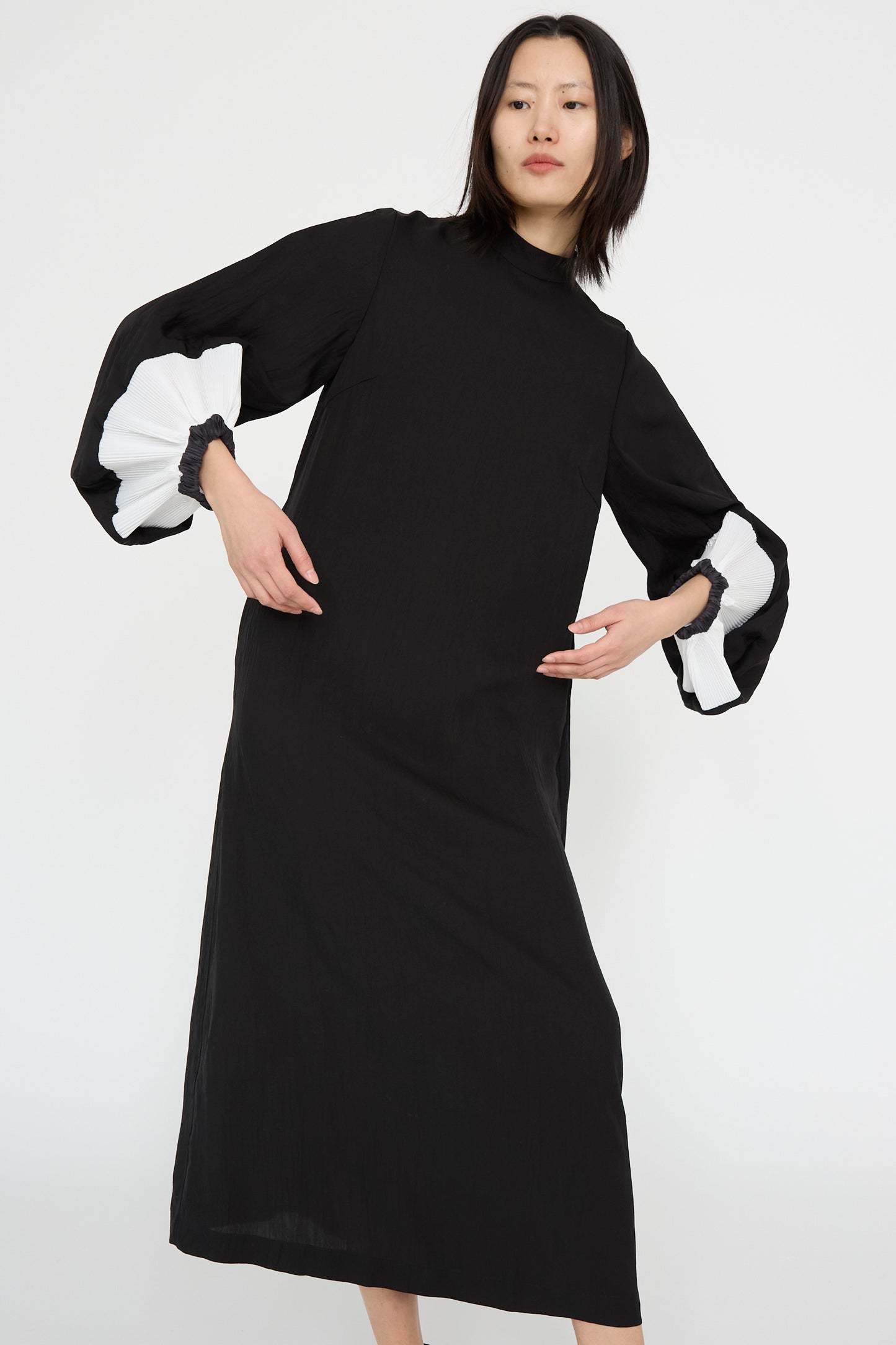 A person poses gracefully in the Acetate Twill Pleats Dress in Black by TOGA ARCHIVES, featuring long bishop sleeves and white accents, against a plain white background.