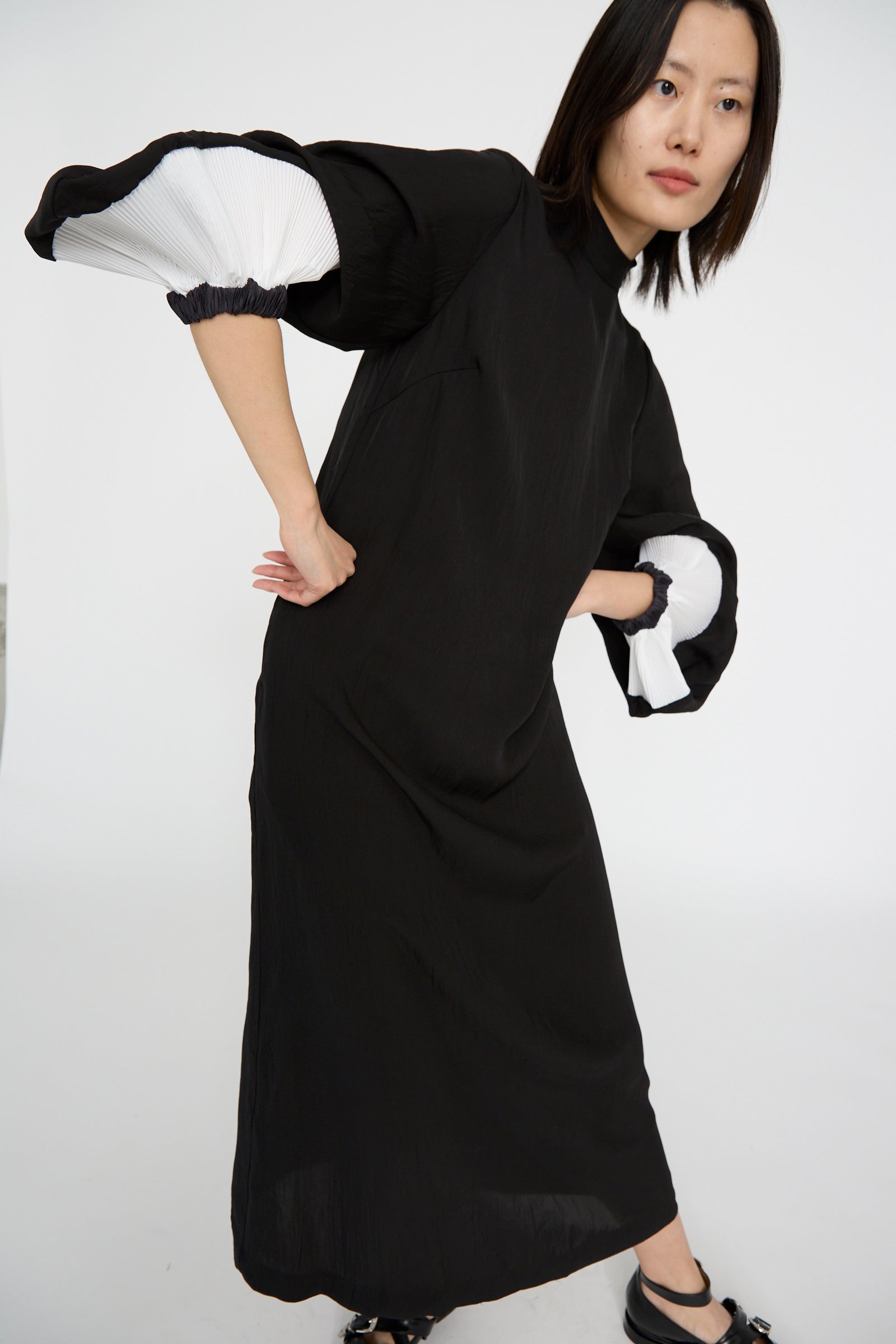 A person in a TOGA ARCHIVES Acetate Twill Pleats Dress in Black, with white pleated bishop sleeves, leans slightly forward against a plain white background.