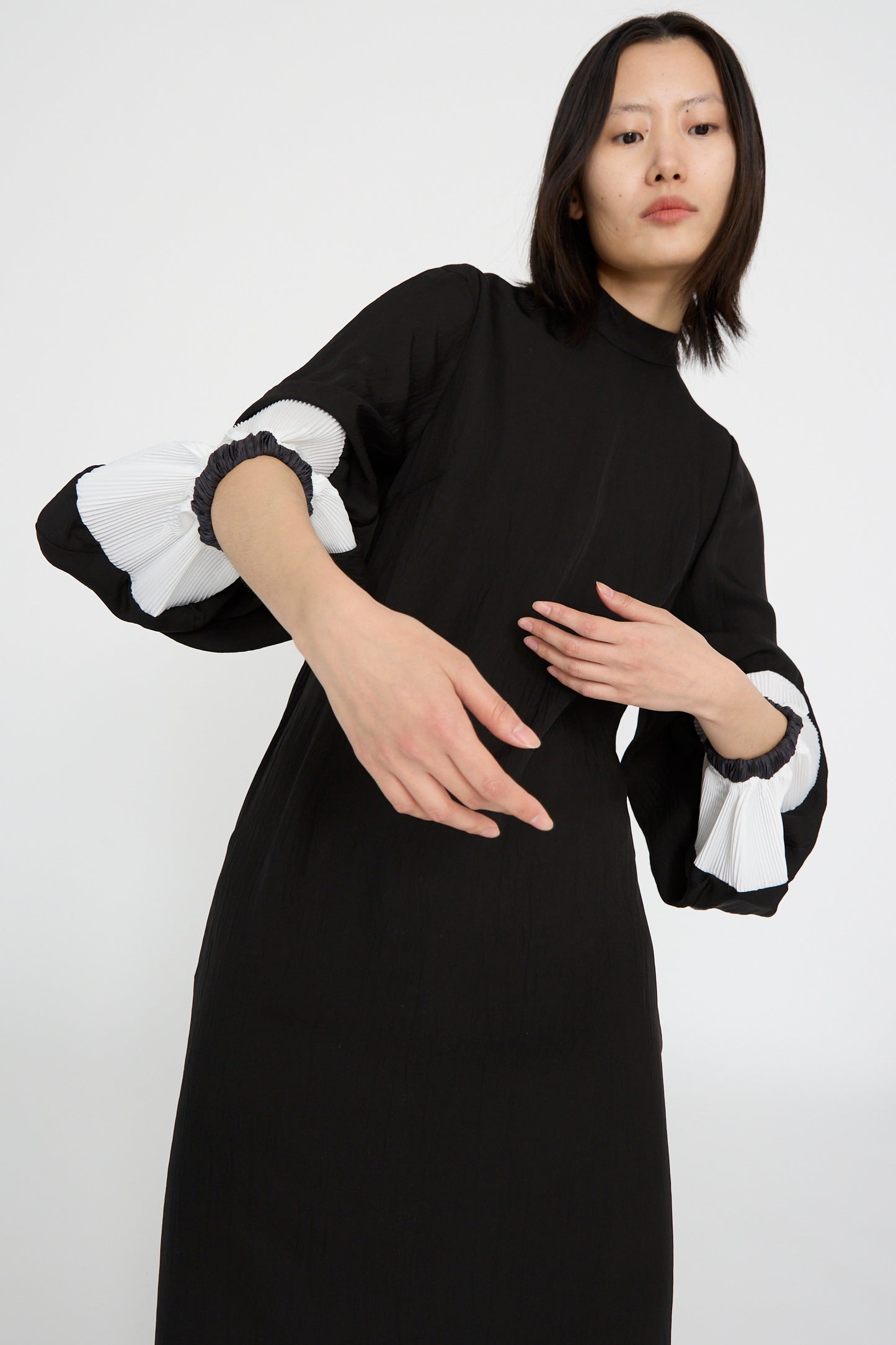 A person stands against a plain white background wearing the TOGA ARCHIVES Acetate Twill Pleats Dress in Black, featuring white ruffled bishop sleeves. Their arms are bent with one hand near the waist and they maintain a neutral expression.