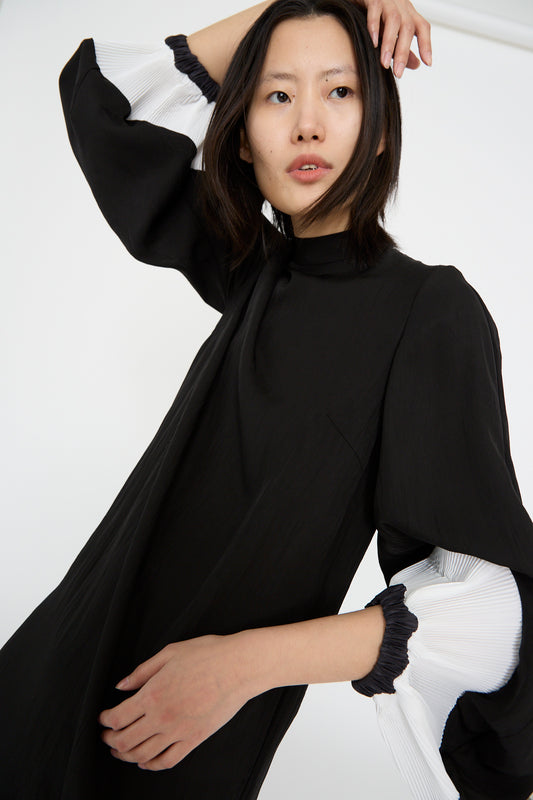 Wearing the Acetate Twill Pleats Dress in Black by TOGA ARCHIVES, a model stands against a white background, looking upwards with one arm raised. The dress's white bishop sleeves create contrast.