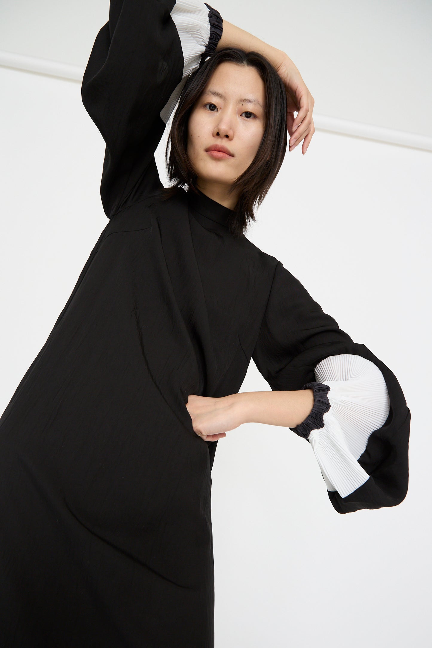 A person poses against a plain backdrop in TOGA ARCHIVES' Acetate Twill Pleats Dress in Black, featuring white cuffs. One hand rests on their hip while the other is raised ad they look forward. 