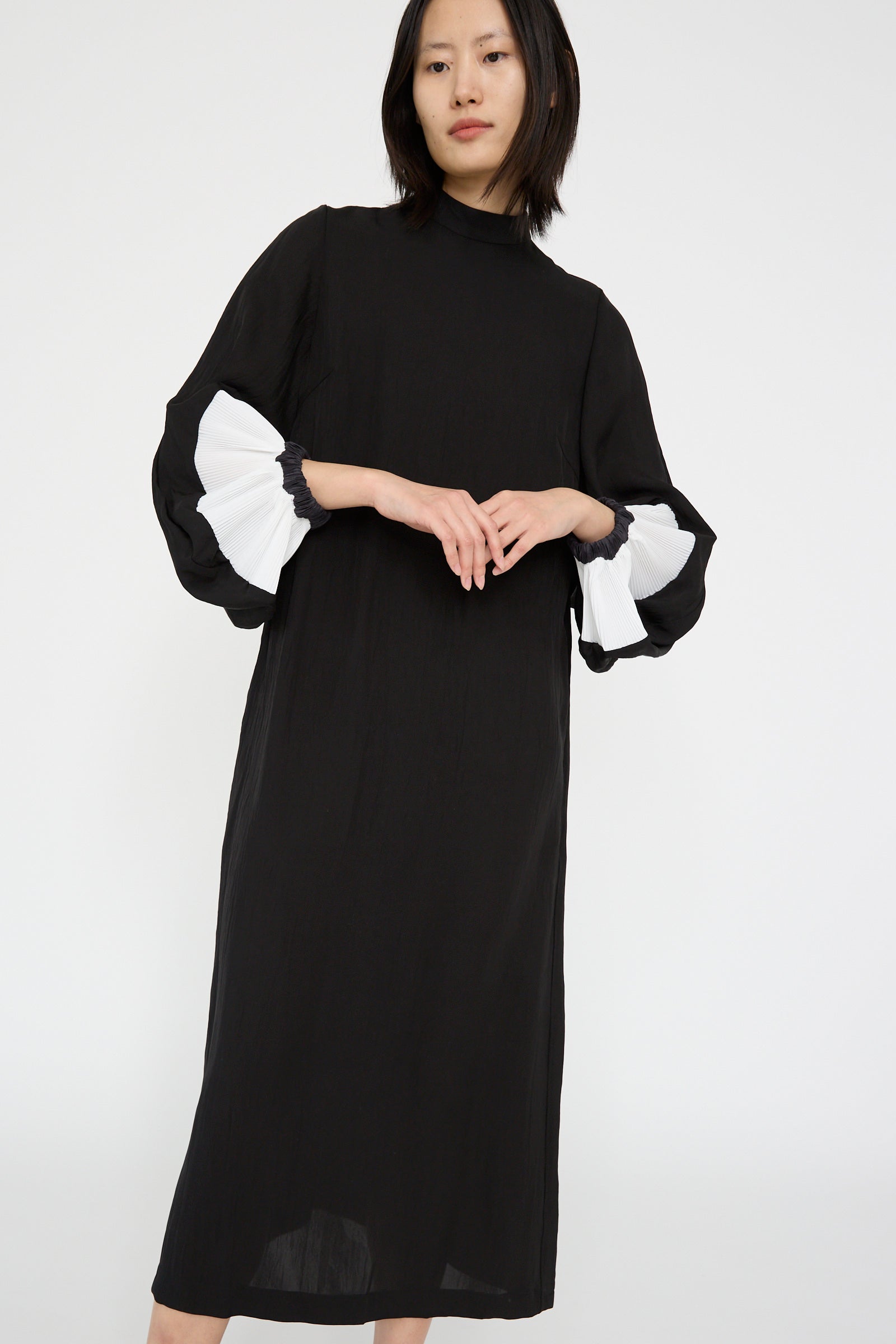 A person stands against a plain white background, dressed in the TOGA ARCHIVES Acetate Twill Pleats Dress in Black, featuring long sleeves and white pleated cuffs, with their hands at their front. 