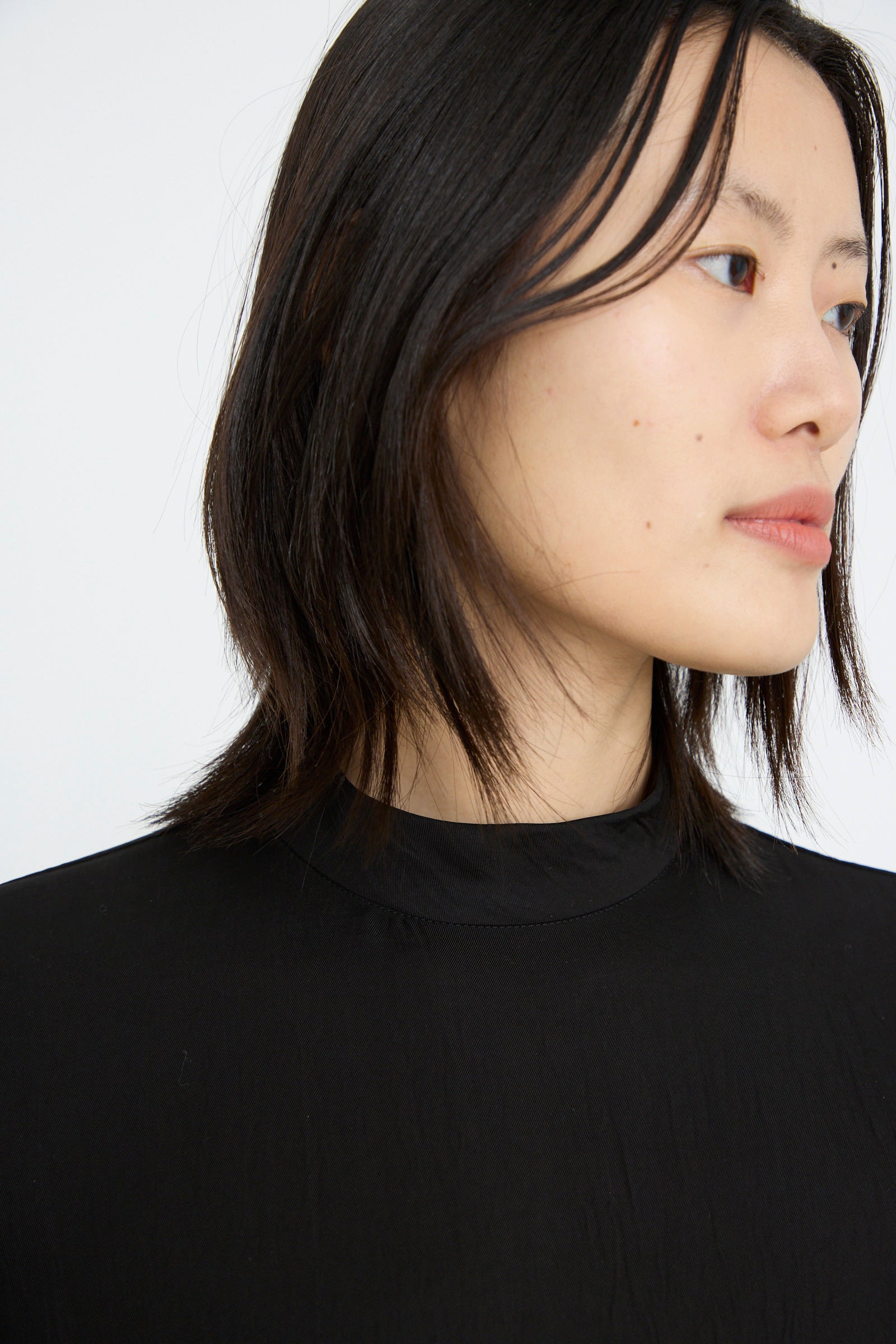 A close-up of a person with shoulder-length black hair wears TOGA ARCHIVES' Acetate Twill Pleats Dress, gazes to the side against a plain background.