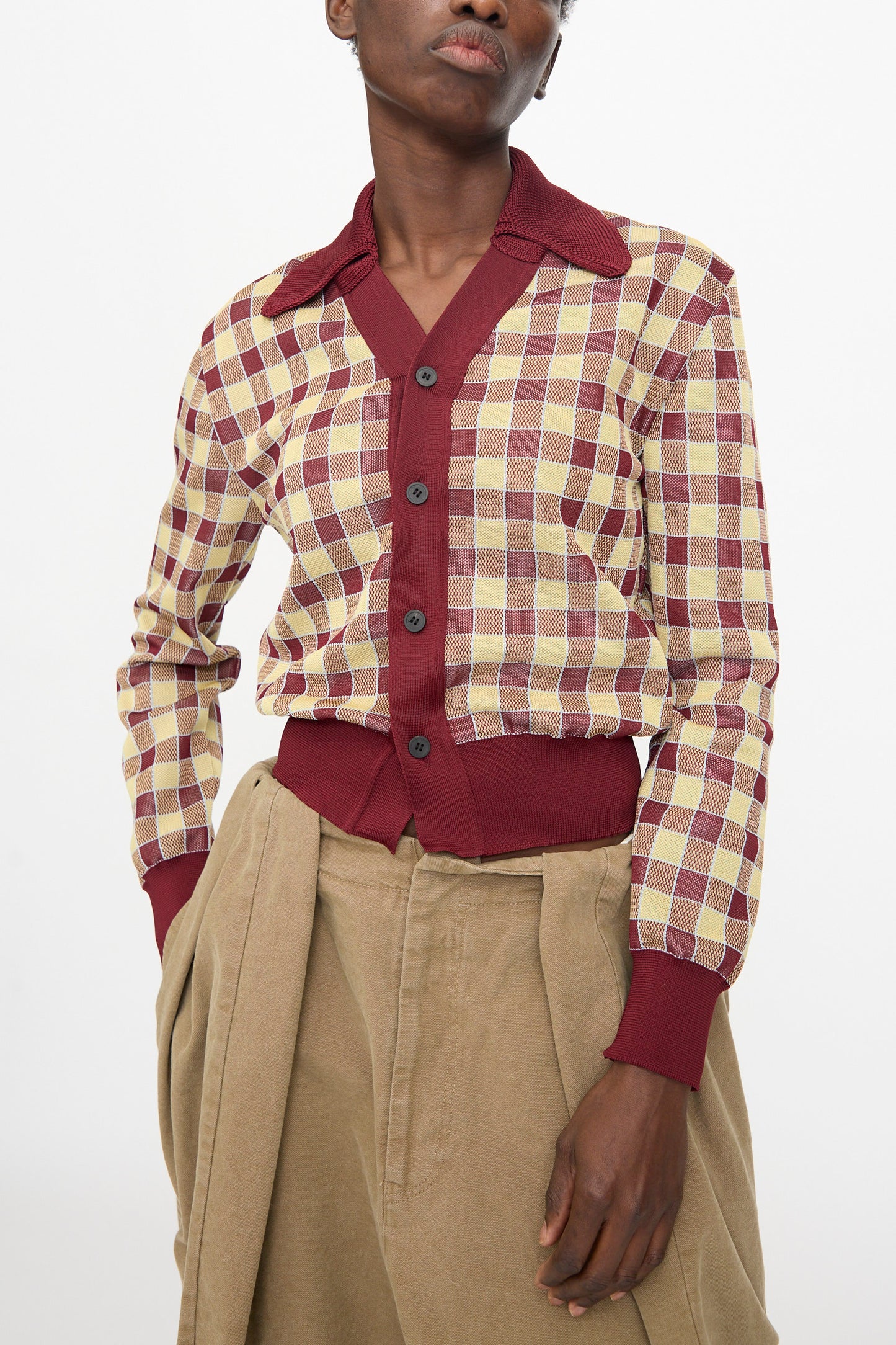 A person wears the TOGA ARCHIVES Check Knit Cardigan in Dark Red, featuring dark buttons and burgundy trim, paired with high-waisted beige pants.
