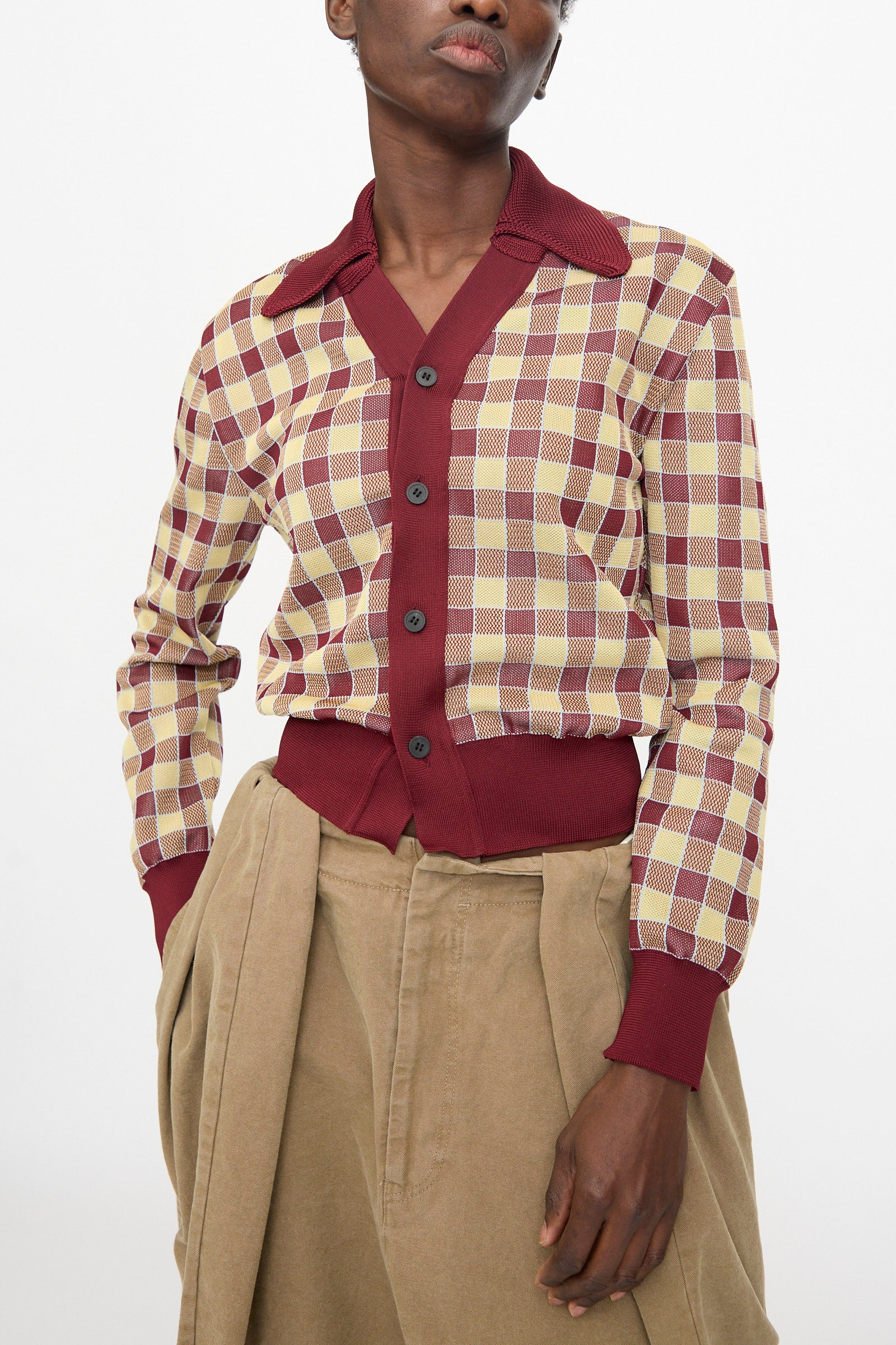 A person wears the TOGA ARCHIVES Check Knit Cardigan in Dark Red, featuring dark buttons and burgundy trim, paired with high-waisted beige pants.