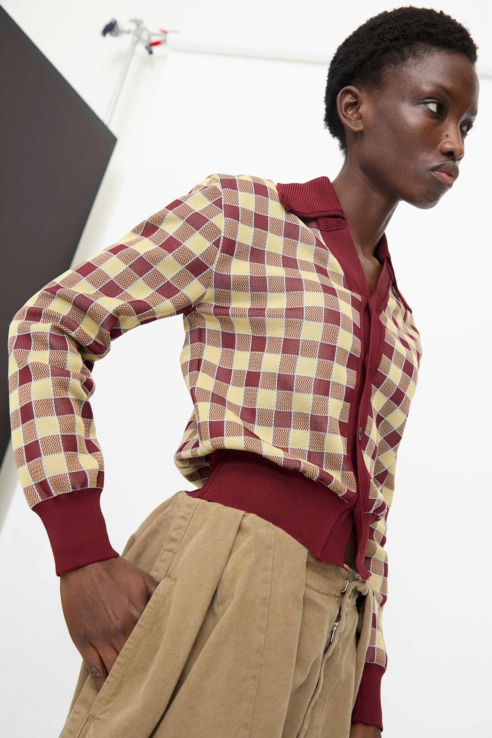A person wears the TOGA ARCHIVES Check Knit Cardigan in Dark Red, paired with beige high-waisted pants, standing against a plain background.