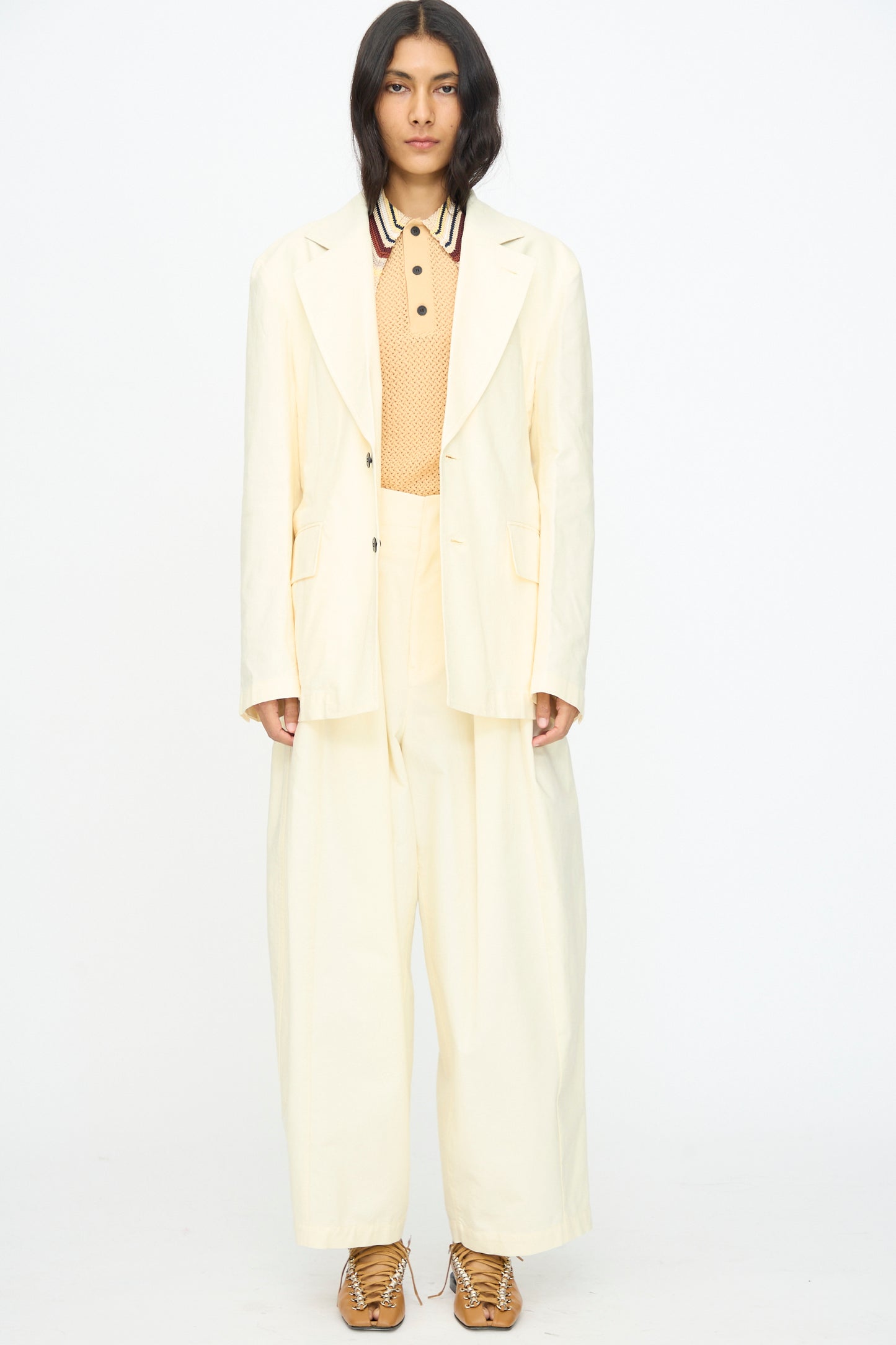 A person stands against a white background wearing the TOGA ARCHIVES Cotton Ox Jacket in Off White, paired with wide-leg trousers, a beige collared shirt, and tan shoes.
