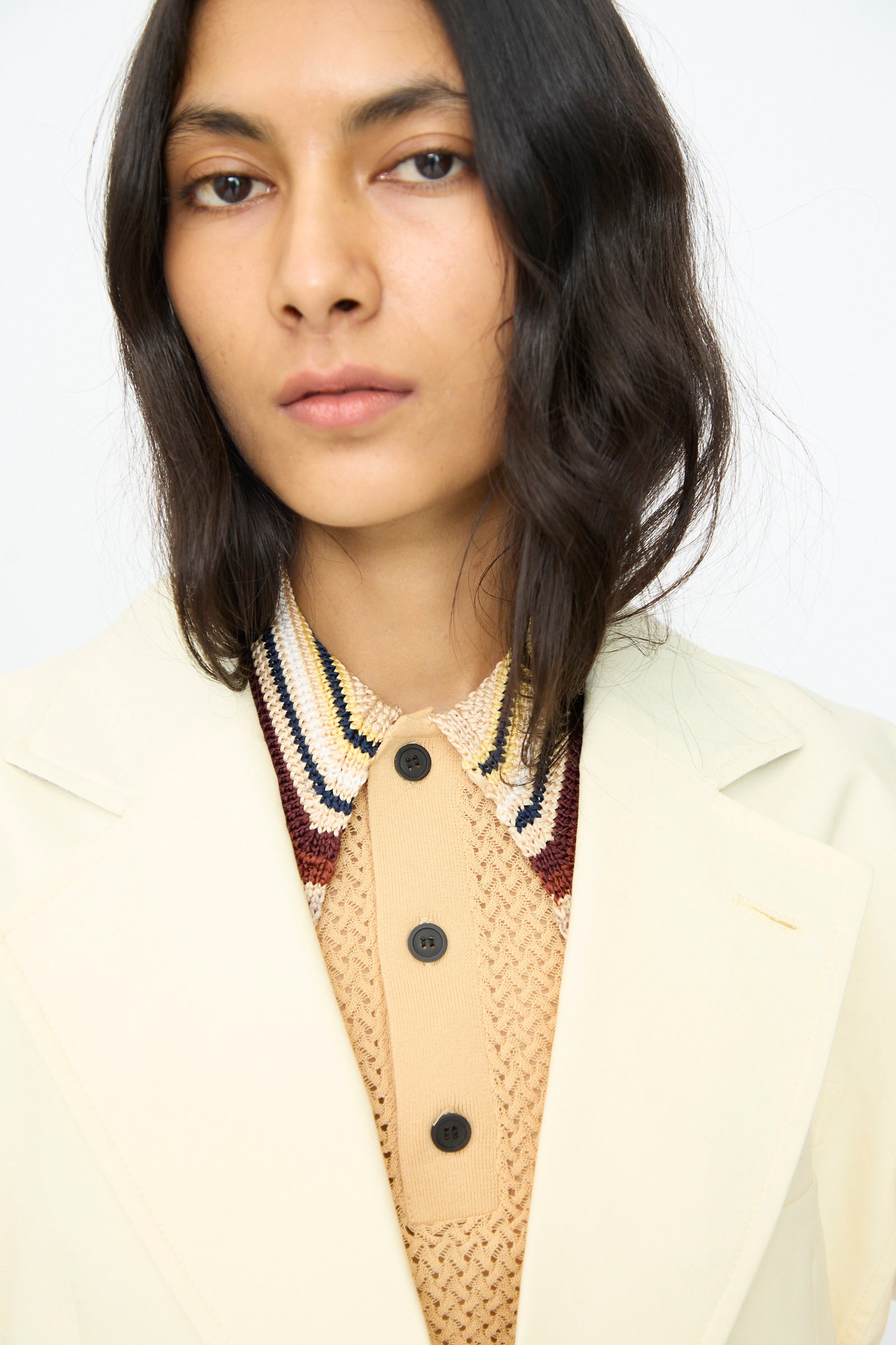 Someone with wavy shoulder-length dark hair wears the TOGA ARCHIVES Cotton Ox Jacket in Off White, made in Japan, layered over a textured beige shirt featuring black buttons and a multicolored collar.