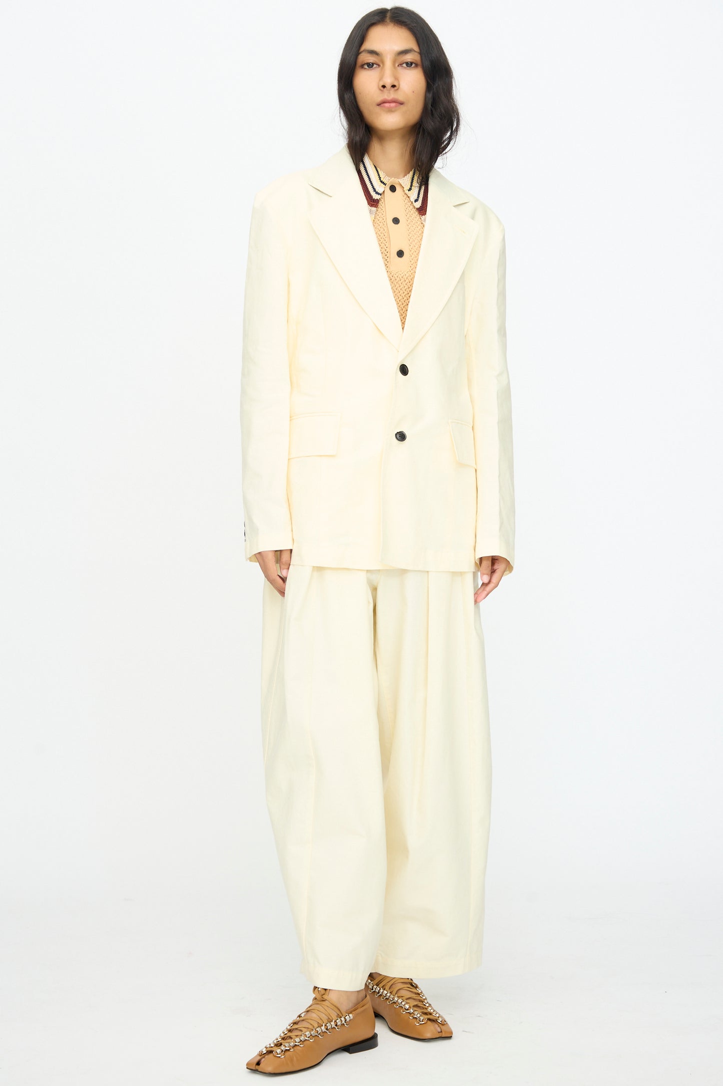 A person stands against a white background wearing the TOGA ARCHIVES Cotton Ox Jacket in Off White, paired with a tan shirt featuring a striped collar and tan lace-up shoes.