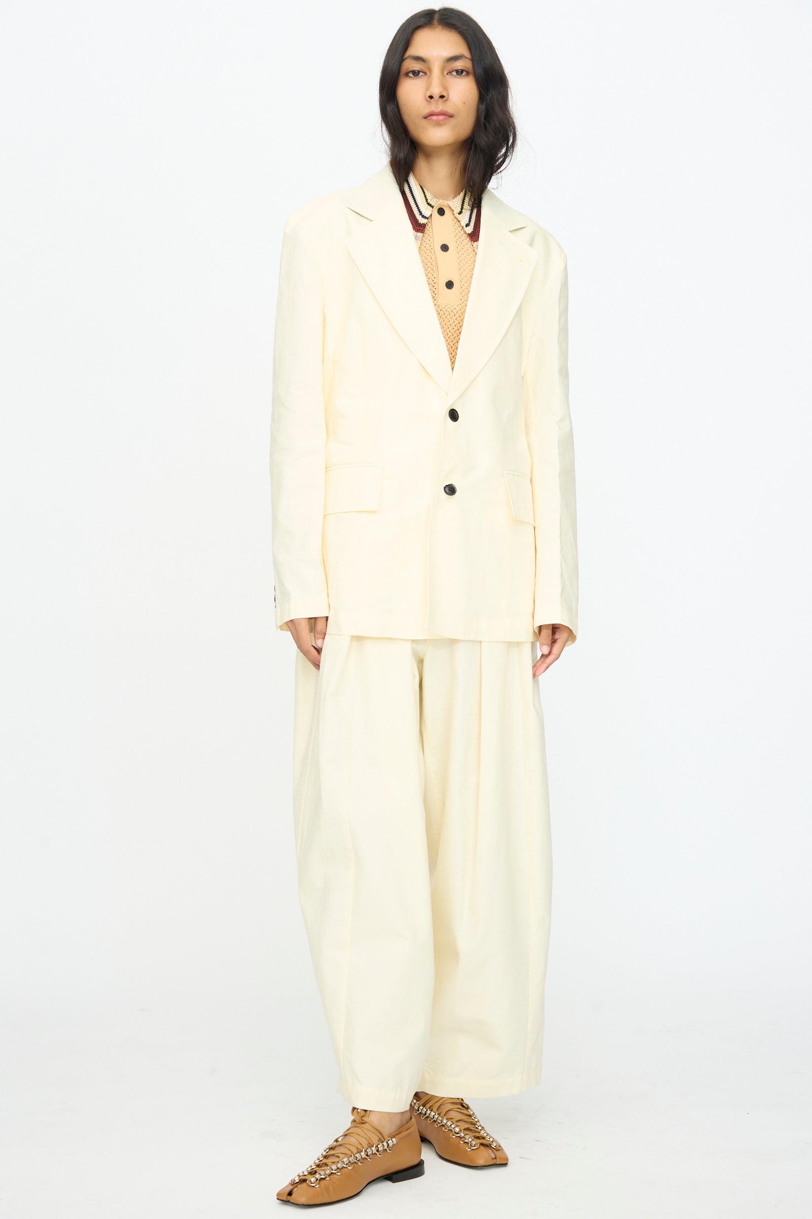 A person stands against a white background wearing the TOGA ARCHIVES Cotton Ox Jacket in Off White, paired with a tan shirt featuring a striped collar and tan lace-up shoes.