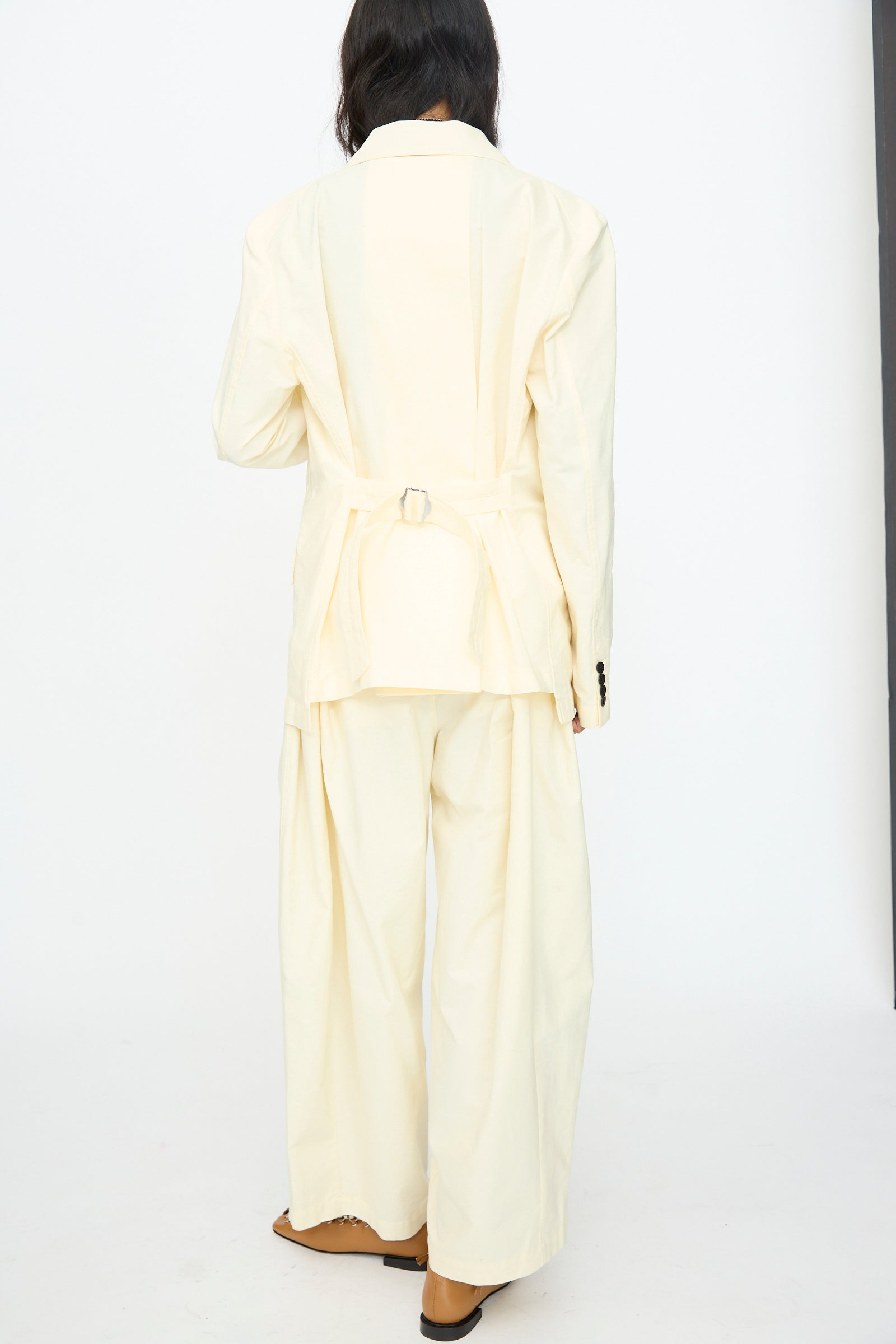 A person stands with their back to the camera, wearing the TOGA ARCHIVES Cotton Ox Jacket in Off White, paired with wide-leg pants and a belted blazer.