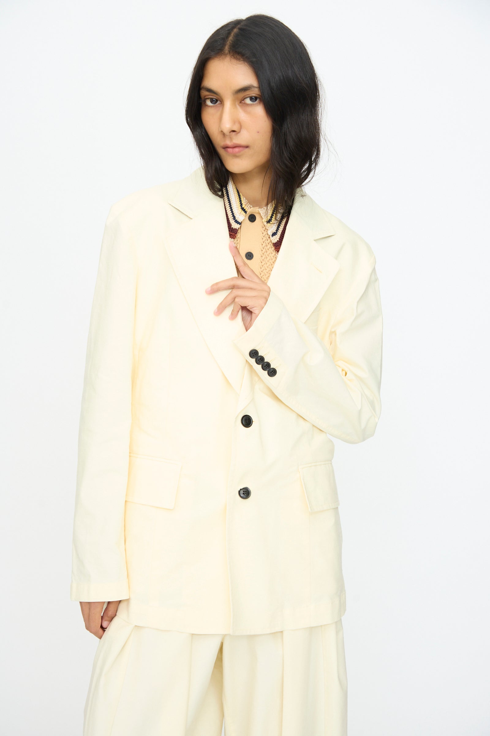 A person wears the TOGA ARCHIVES Cotton Ox Jacket in Off White with black buttons and wide-leg pants, standing against a plain white background and touching the lapel. This relaxed jacket is expertly crafted in Japan.