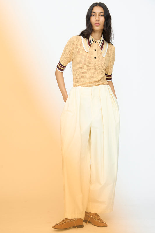 A woman stands against a light background wearing the TOGA ARCHIVES Cotton Ox Wide Pant in Off White, paired with a beige short-sleeve collared striped shirt and beige woven shoes.