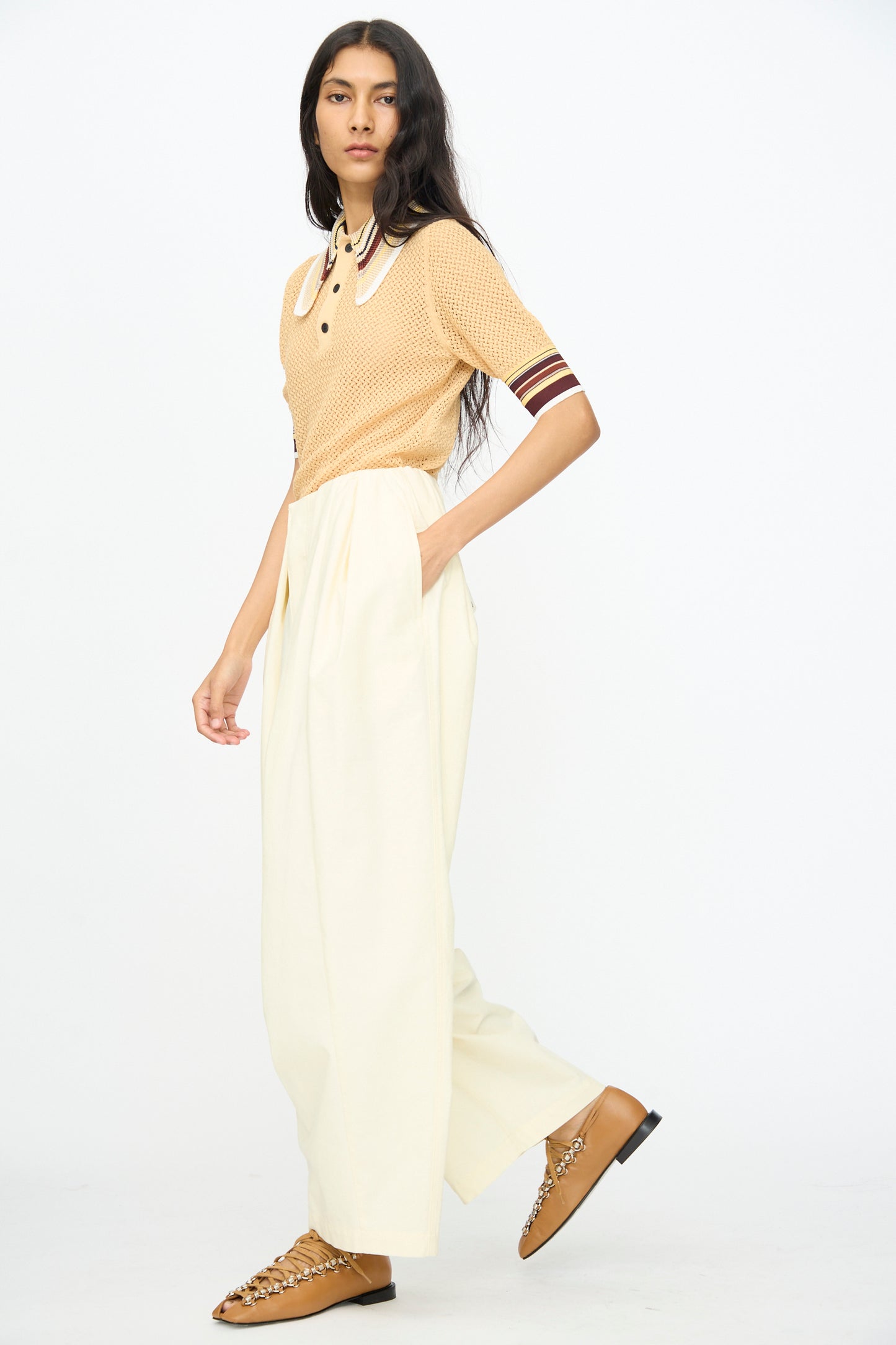 A person stands against a plain background, wearing an off white short-sleeve knit top, TOGA ARCHIVES Cotton Ox Wide Pant in Off White, and tan chain detail loafers.
