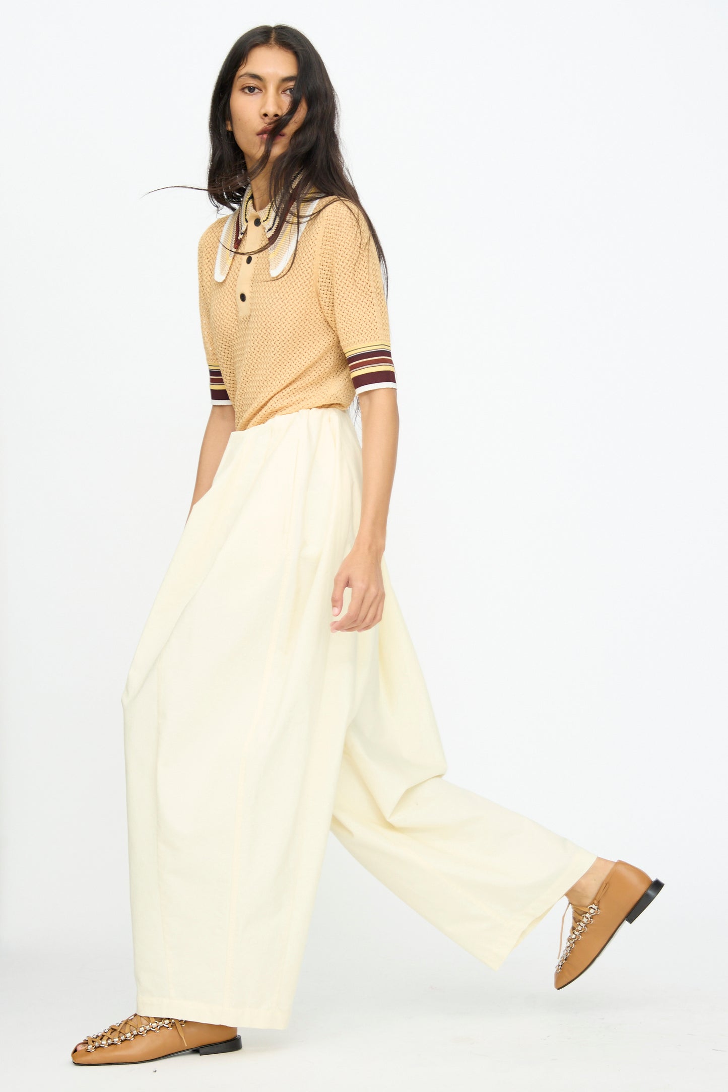 Wearing a tan short-sleeve knit top with a white collar, TOGA ARCHIVES Cotton Ox Wide Pant in Off White, and tan loafers with chain detail, the person stands against a plain white background.