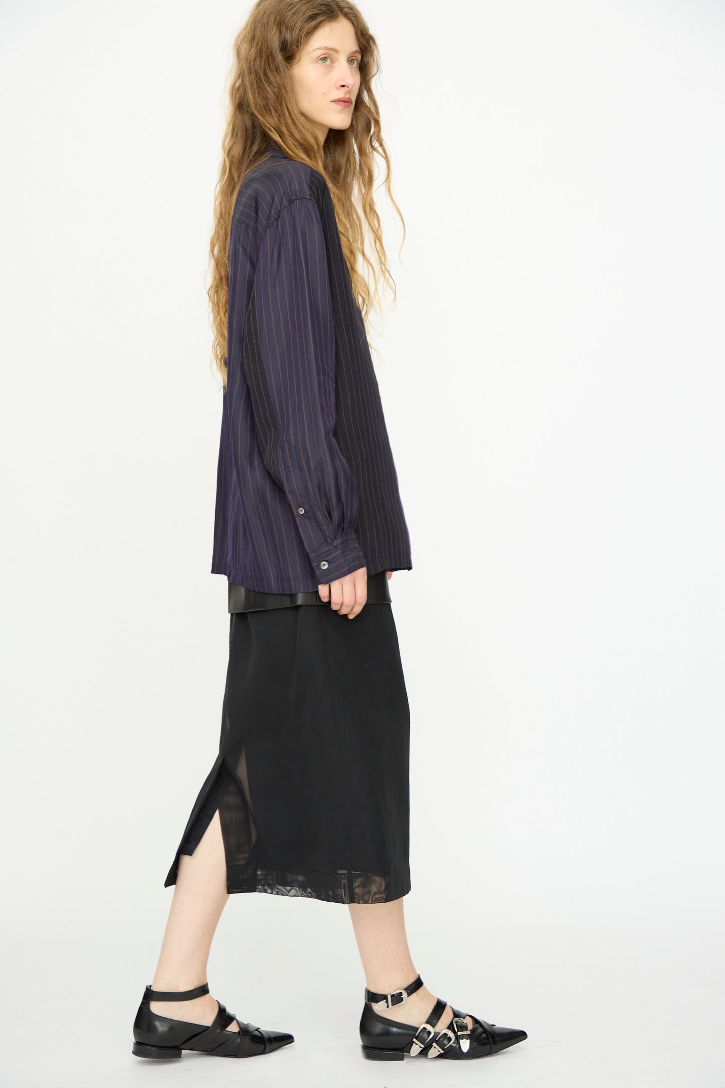 A woman with long wavy hair wears the TOGA ARCHIVES Cupra Stripe Shirt in Navy, paired with a black midi skirt featuring a side slit and black buckled flats, standing against a plain white background.