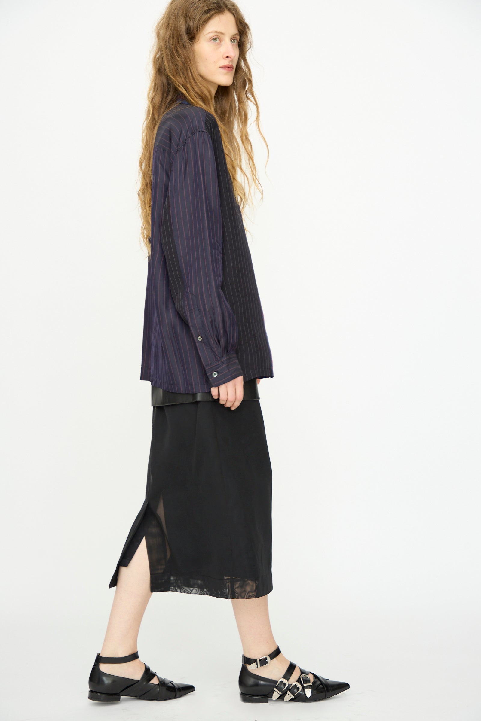 A woman with long wavy hair wears the TOGA ARCHIVES Cupra Stripe Shirt in Navy, paired with a black midi skirt featuring a side slit and black buckled flats, standing against a plain white background.