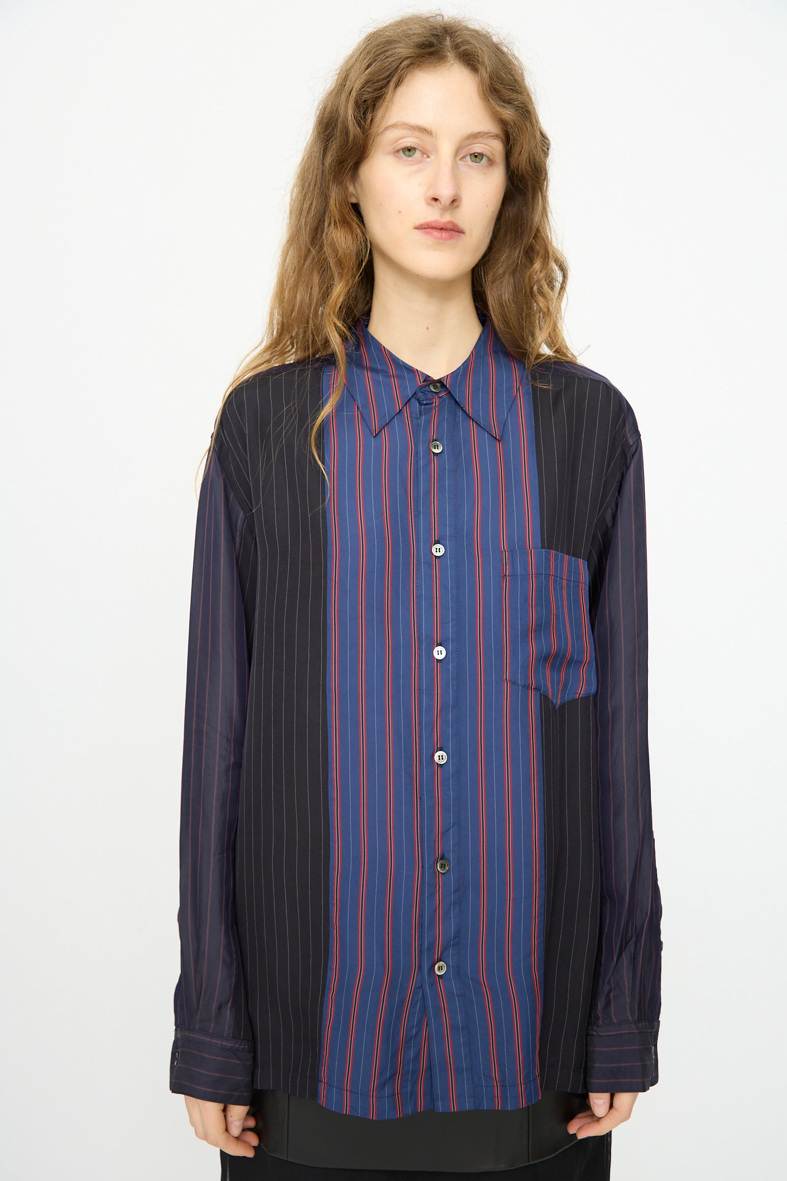 A person with long wavy hair wears the TOGA ARCHIVES Cupra Stripe Shirt in Navy—featuring loose fit, blue and black panels, and red pinstripes—standing against a plain white background.