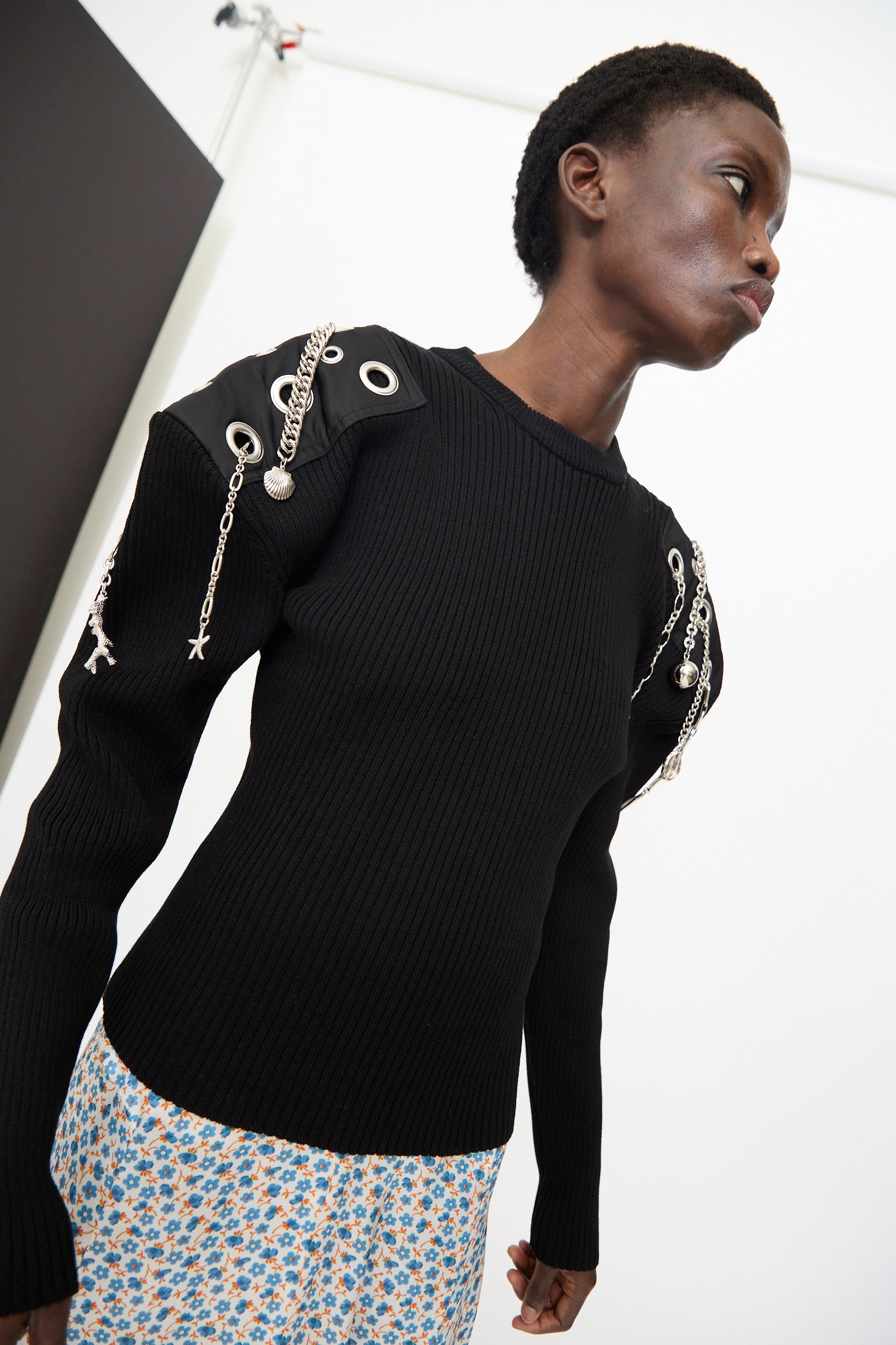 A person wears the TOGA ARCHIVES Decoration Rib Knit Pullover in Black, featuring metal chain and grommet details on the shoulders, paired with a blue floral skirt, standing against a white background.