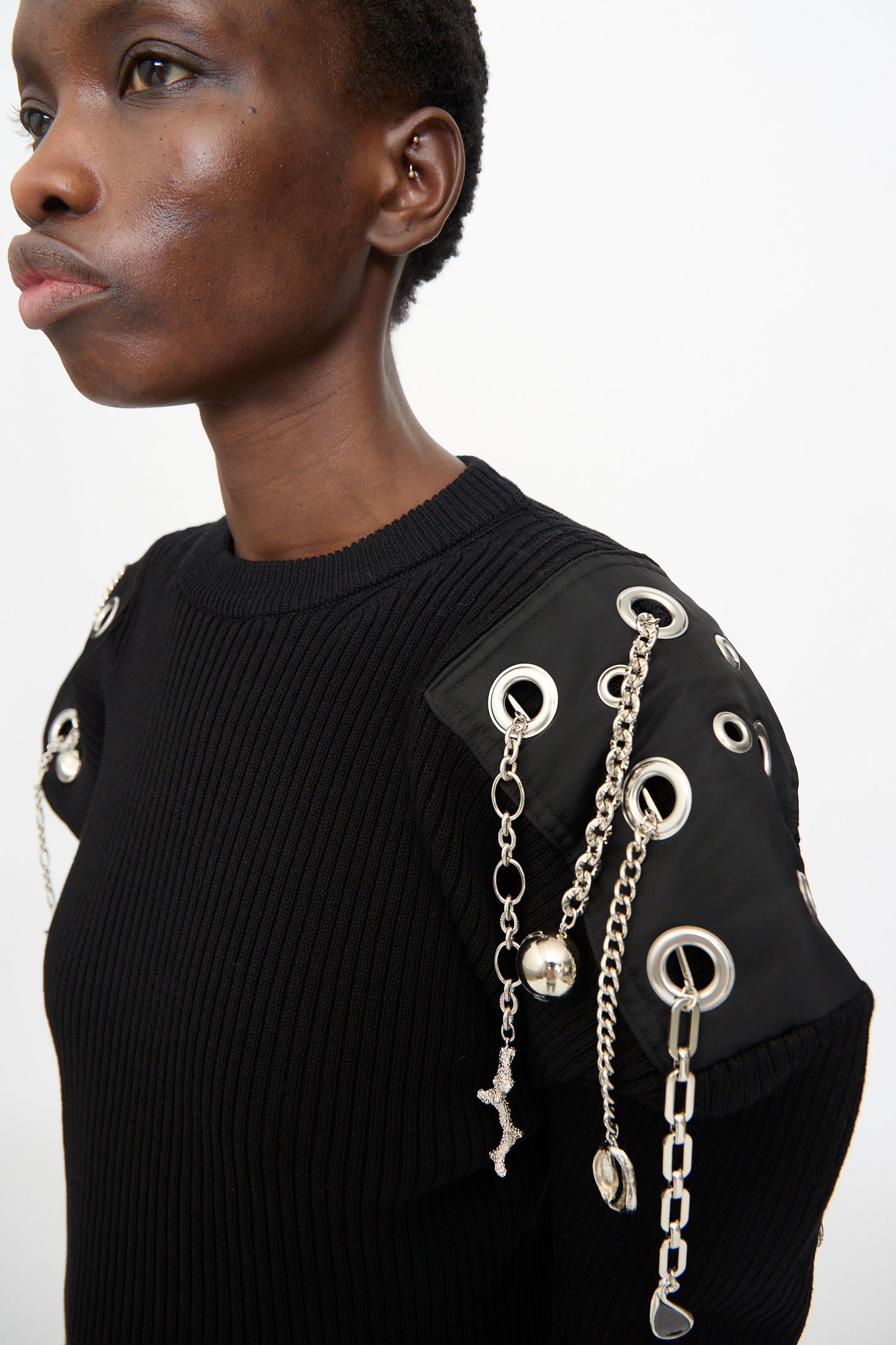 A model wears the TOGA ARCHIVES Decoration Rib Knit Pullover in Black, featuring metallic grommets and hanging silver chains on the shoulders, set against a plain white background.