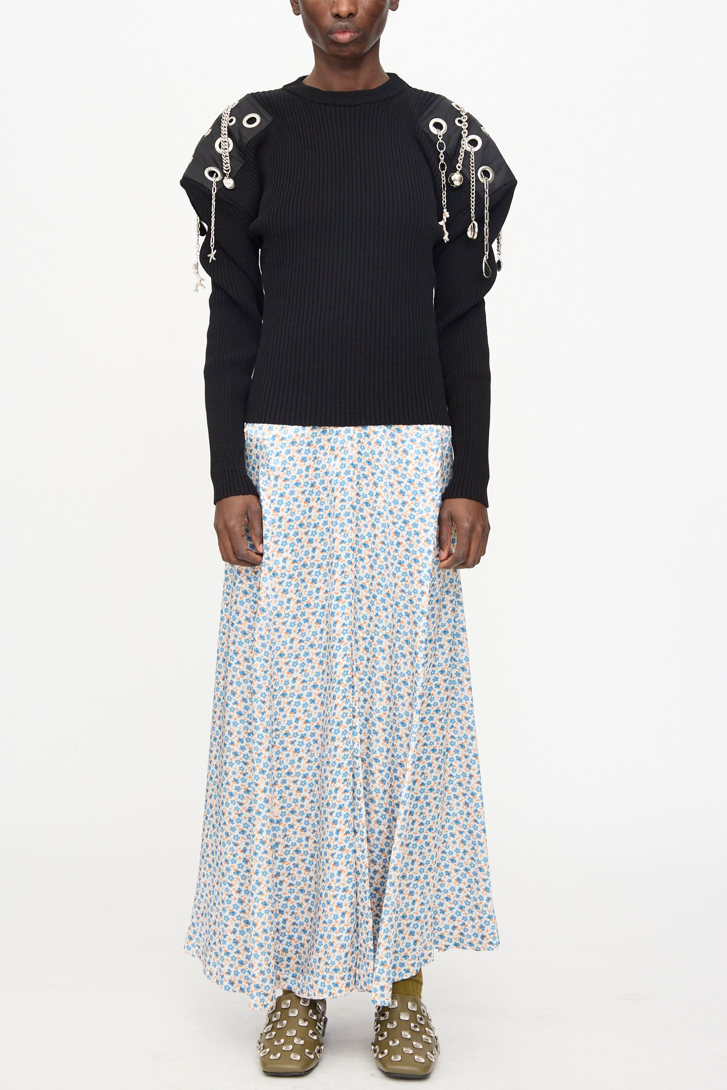 A person wears the TOGA ARCHIVES Decoration Rib Knit Pullover in Black, paired with a long light blue floral skirt and olive green shoes with white embellishments, standing against a plain background.