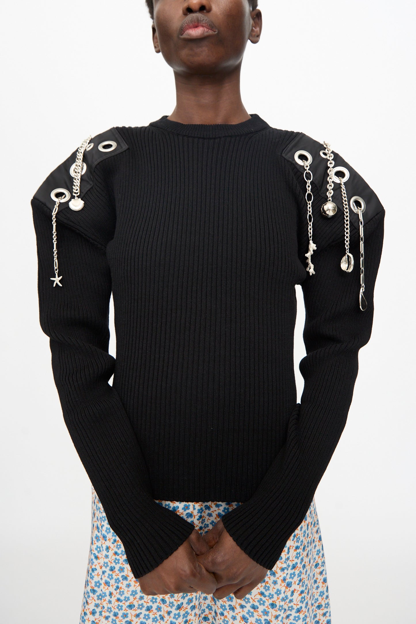 A person wears the TOGA ARCHIVES Decoration Rib Knit Pullover in Black, featuring metallic grommets and shoulder chains, paired with a blue floral skirt against a plain background.