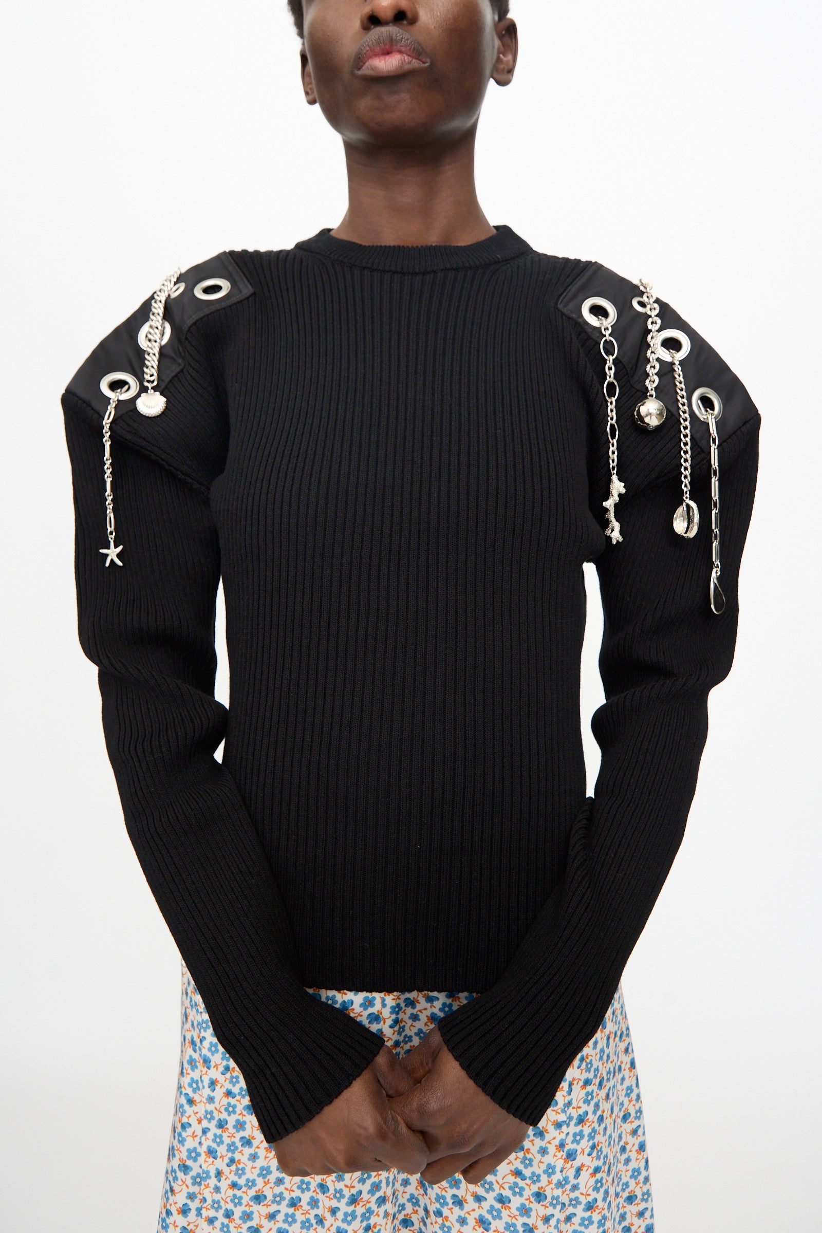 A person wears the TOGA ARCHIVES Decoration Rib Knit Pullover in Black, featuring metallic grommets and shoulder chains, paired with a blue floral skirt against a plain background.